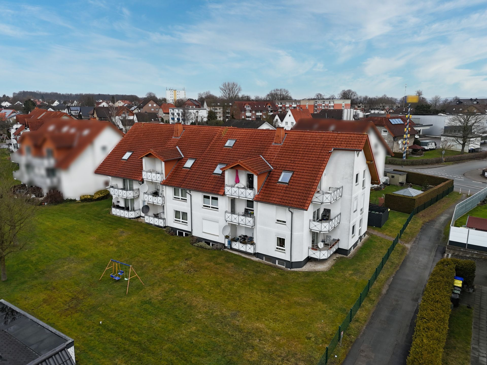 Mehrfamilienhaus mit rotem Dach, Balkonen und großem Garten auf einer Wiese.