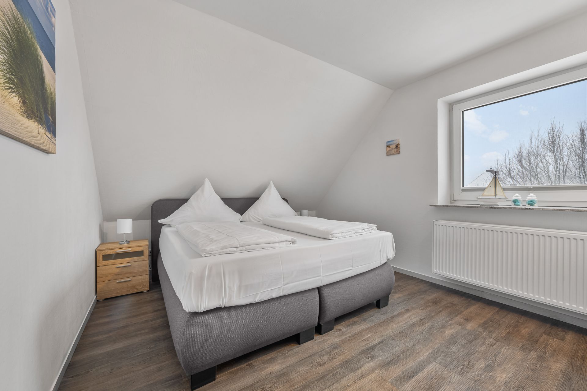 Helles Schlafzimmer mit Doppelbett, Nachttisch, Fenster und Holzfußboden.