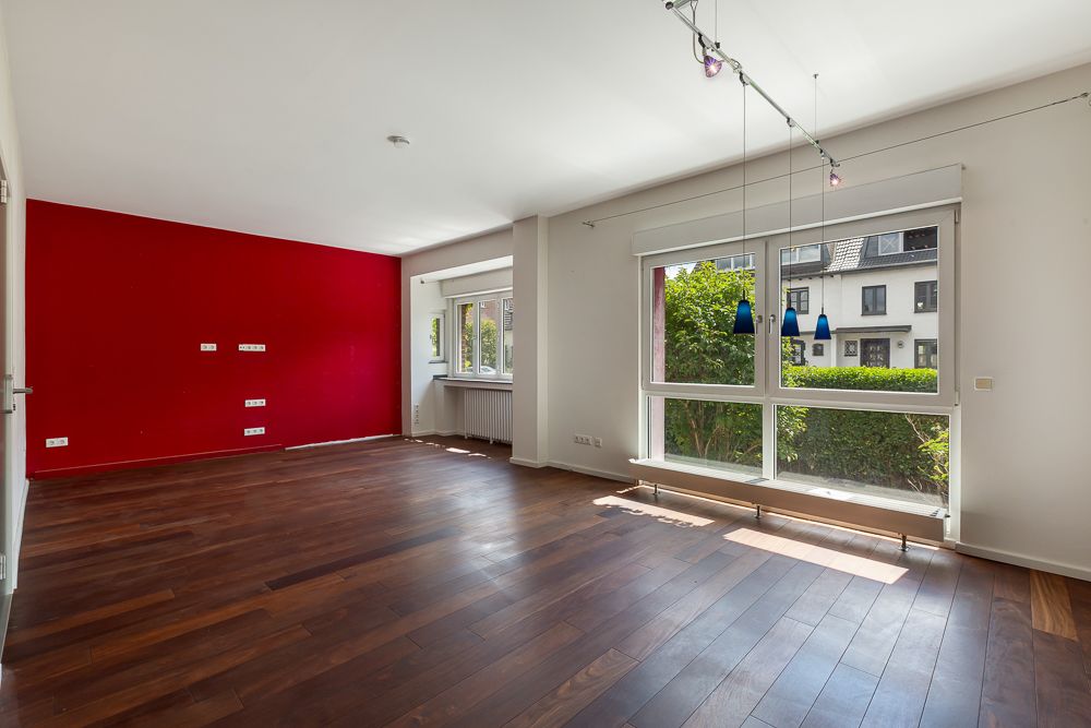 Heller Wohnraum mit Holzfußboden, roter Akzentwand und großen Fenstern zur Straße.