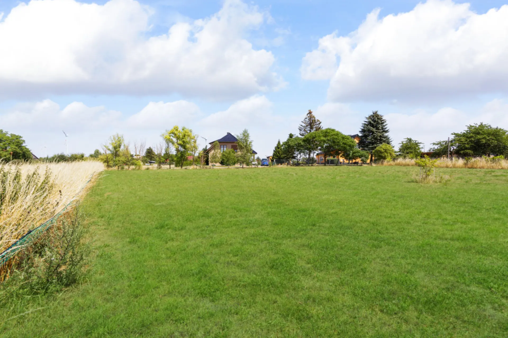 Grünes Grundstück mit Bäumen und Haus im Hintergrund unter blauem Himmel mit Wolken.
