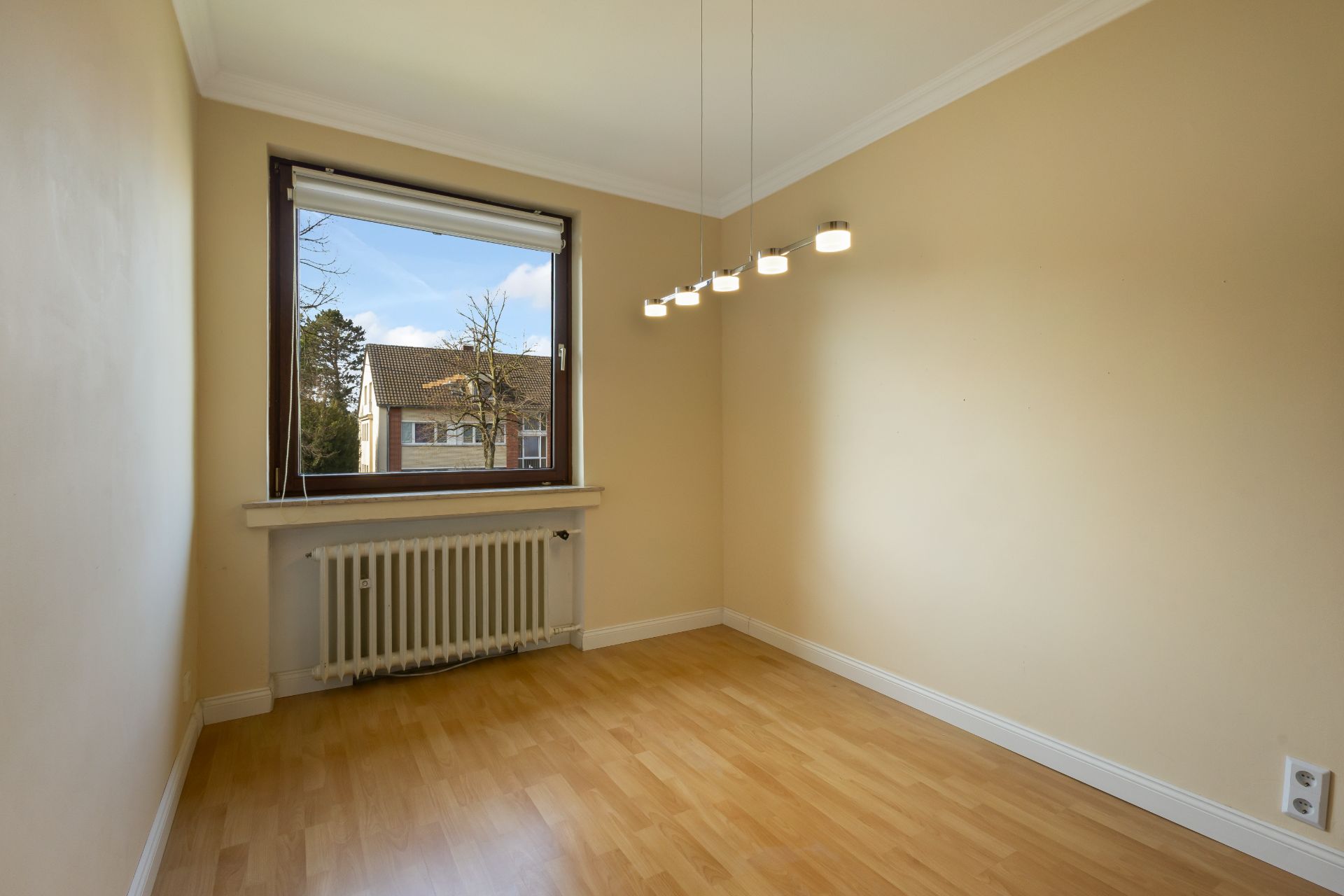 Leerer Raum mit Holzfußboden, großem Fenster, Heizkörper und moderner Deckenleuchte.