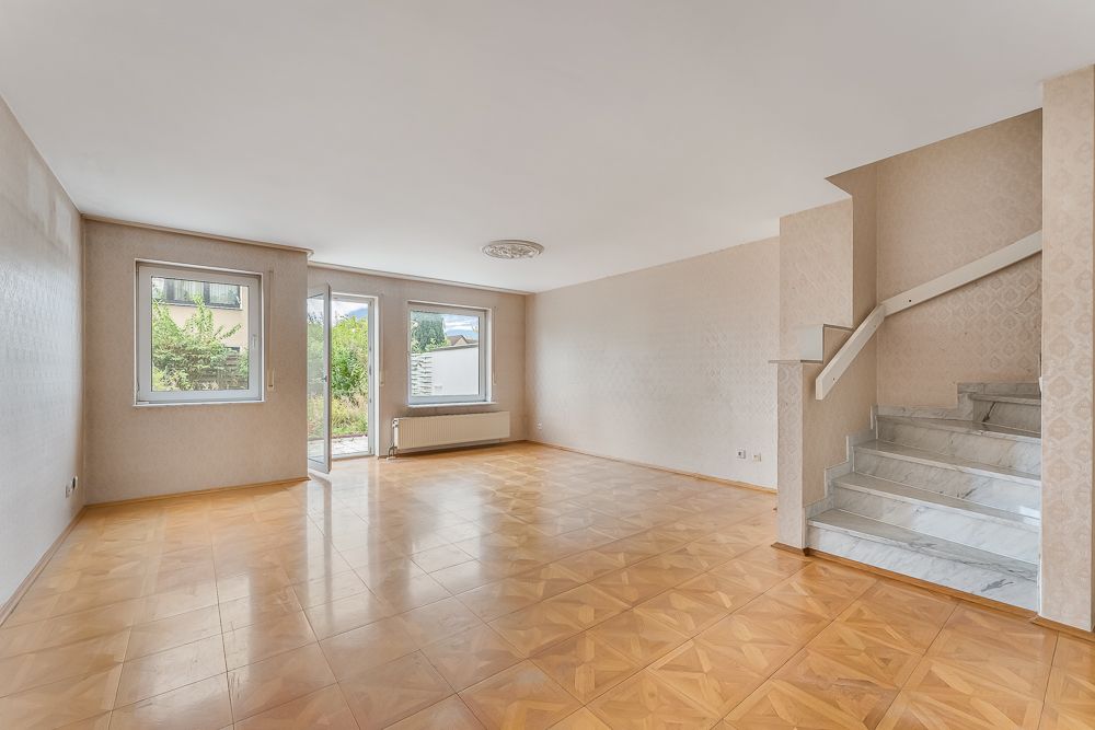 Helles, leeres Wohnzimmer mit Parkettboden, zwei Fenstern, Glastür und Marmortreppe.