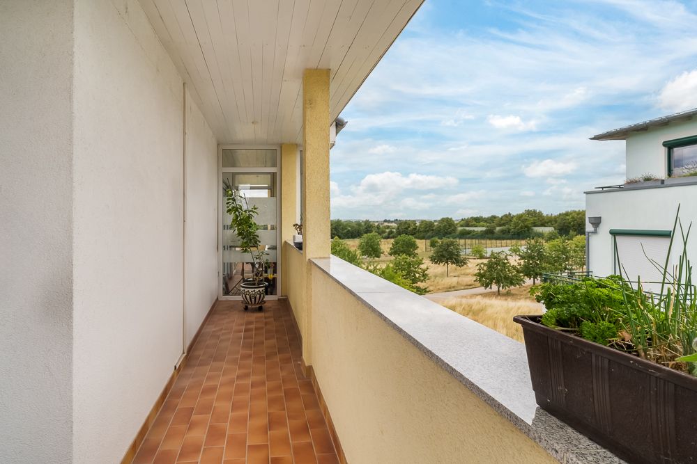 Langer Balkon mit braunen Fliesen und Pflanzen, Blick auf grüne Landschaft.