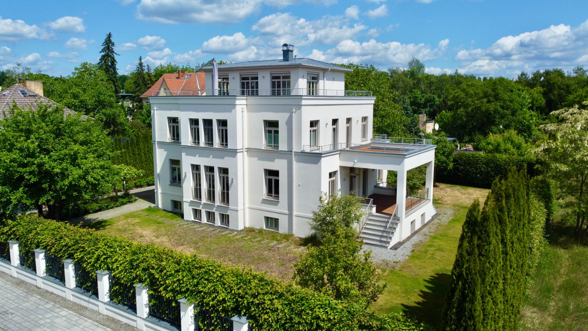 Modernes, weißes Wohnhaus mit drei Etagen, Garten und Terrasse, umgeben von Bäumen und Hecken.