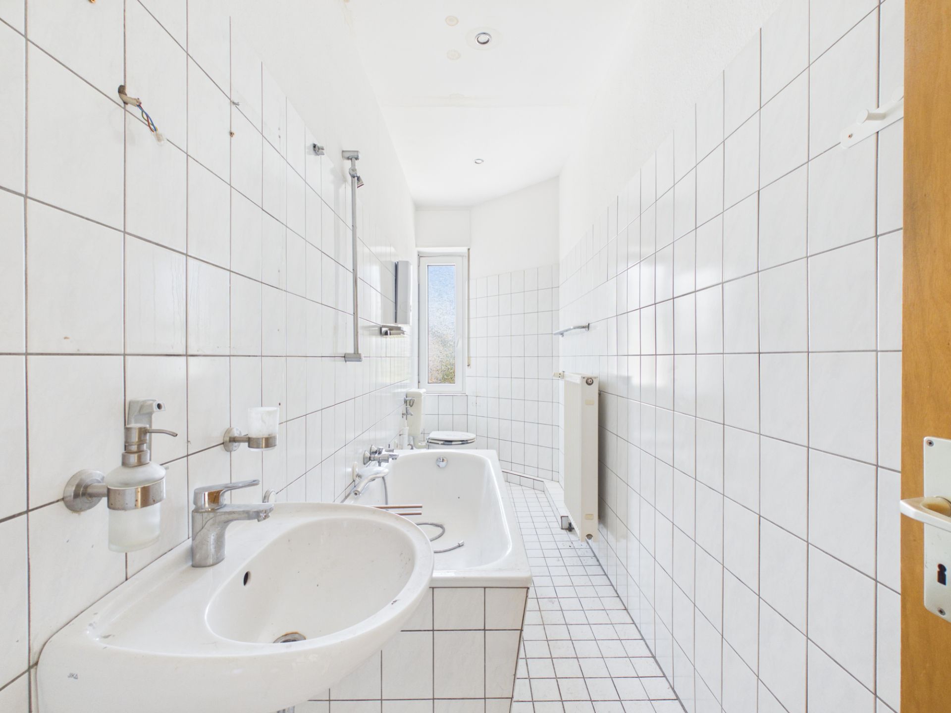 Weiß gefliestes Badezimmer mit Waschbecken, Badewanne und Fenster am Ende des Raumes.