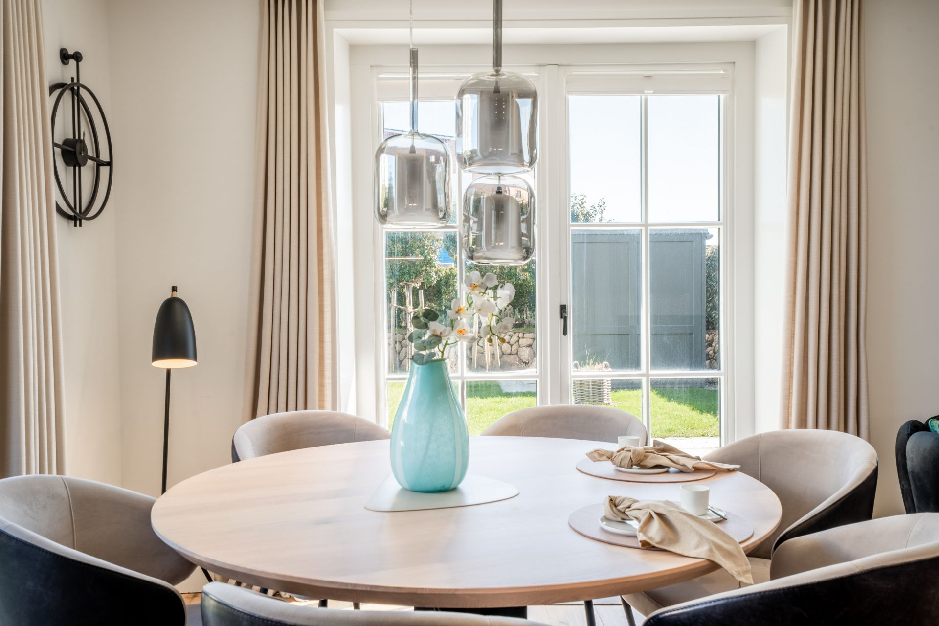 Essbereich mit rundem Holztisch, Vase, vier Stühlen und großer Fensterfront im Hintergrund.