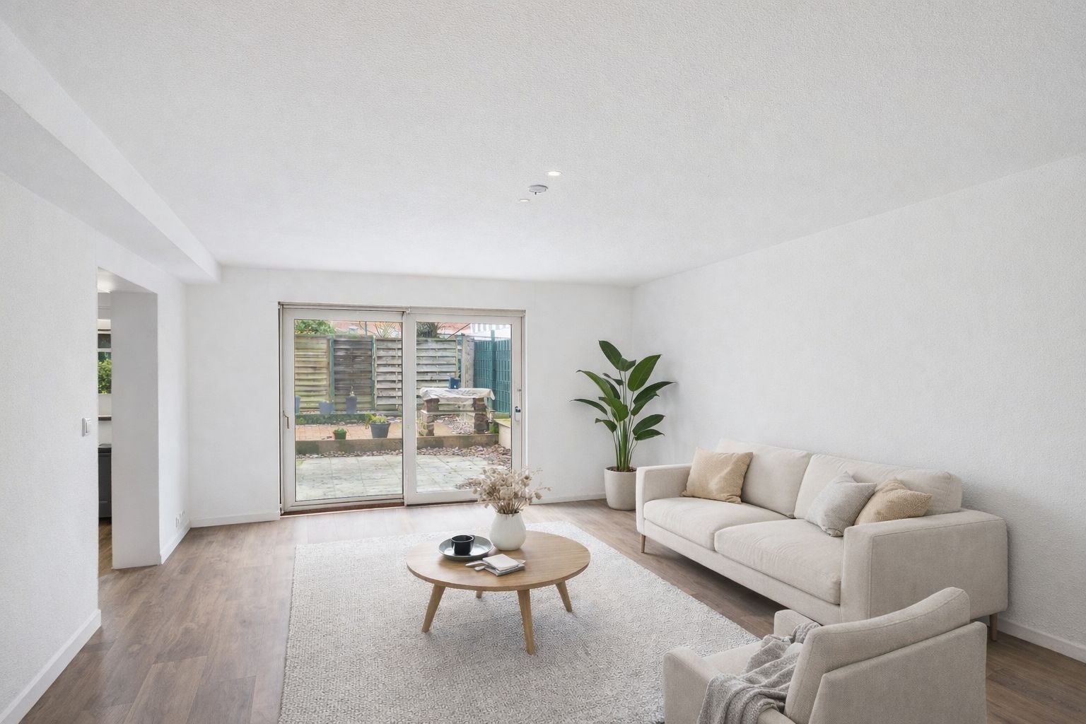 Helles Wohnzimmer mit Sofa, Sessel, rundem Tisch, Teppich und großer Glastür zum Garten.