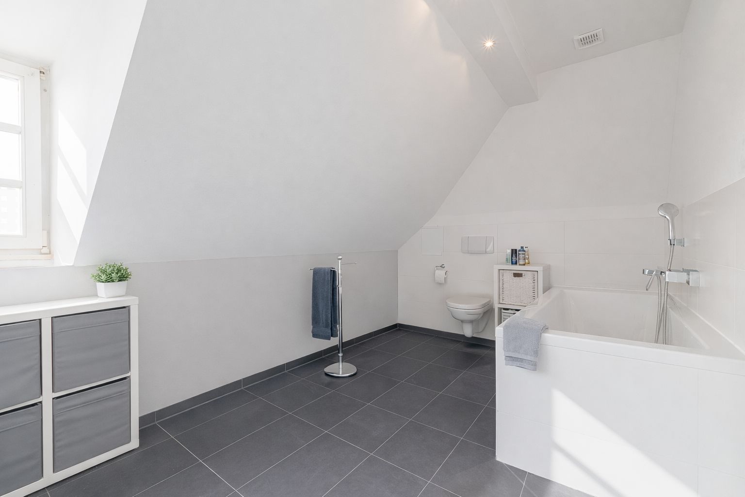 Helles Badezimmer mit Badewanne, WC, Fenster und Fliesenboden unter einer Dachschräge.