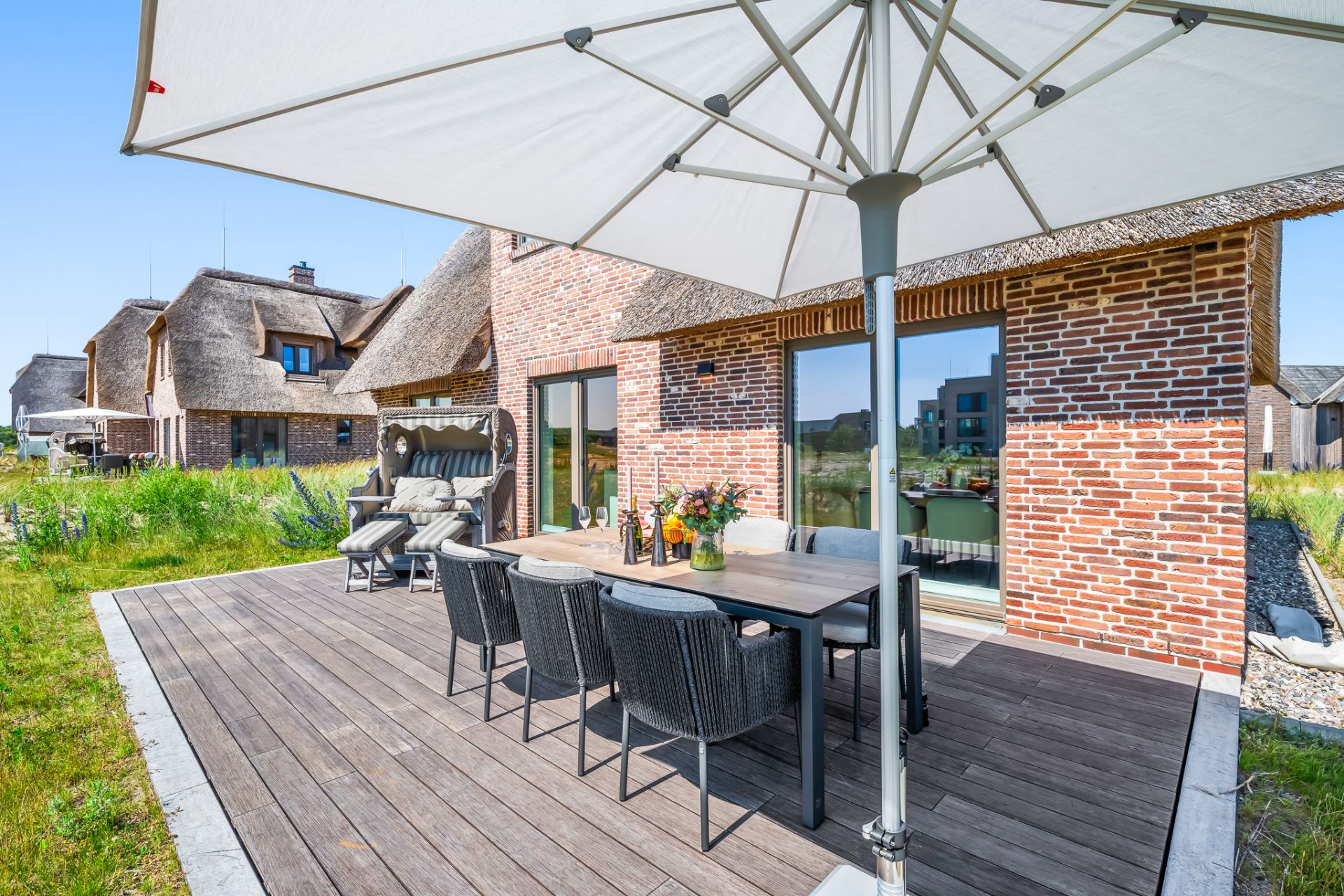 Holzterrasse mit Esstisch, Stühlen und Sonnenschirm vor einem Haus mit Reetdach und Backsteinfassade.