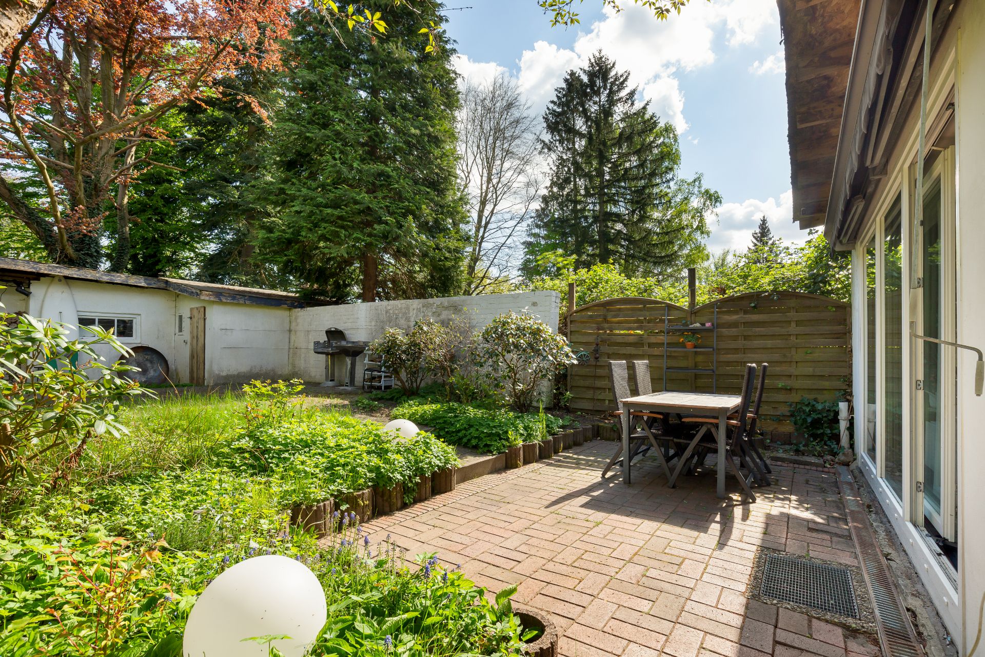 Gepflasterte Terrasse mit Tisch und Stühlen, umgeben von Garten und Bäumen.