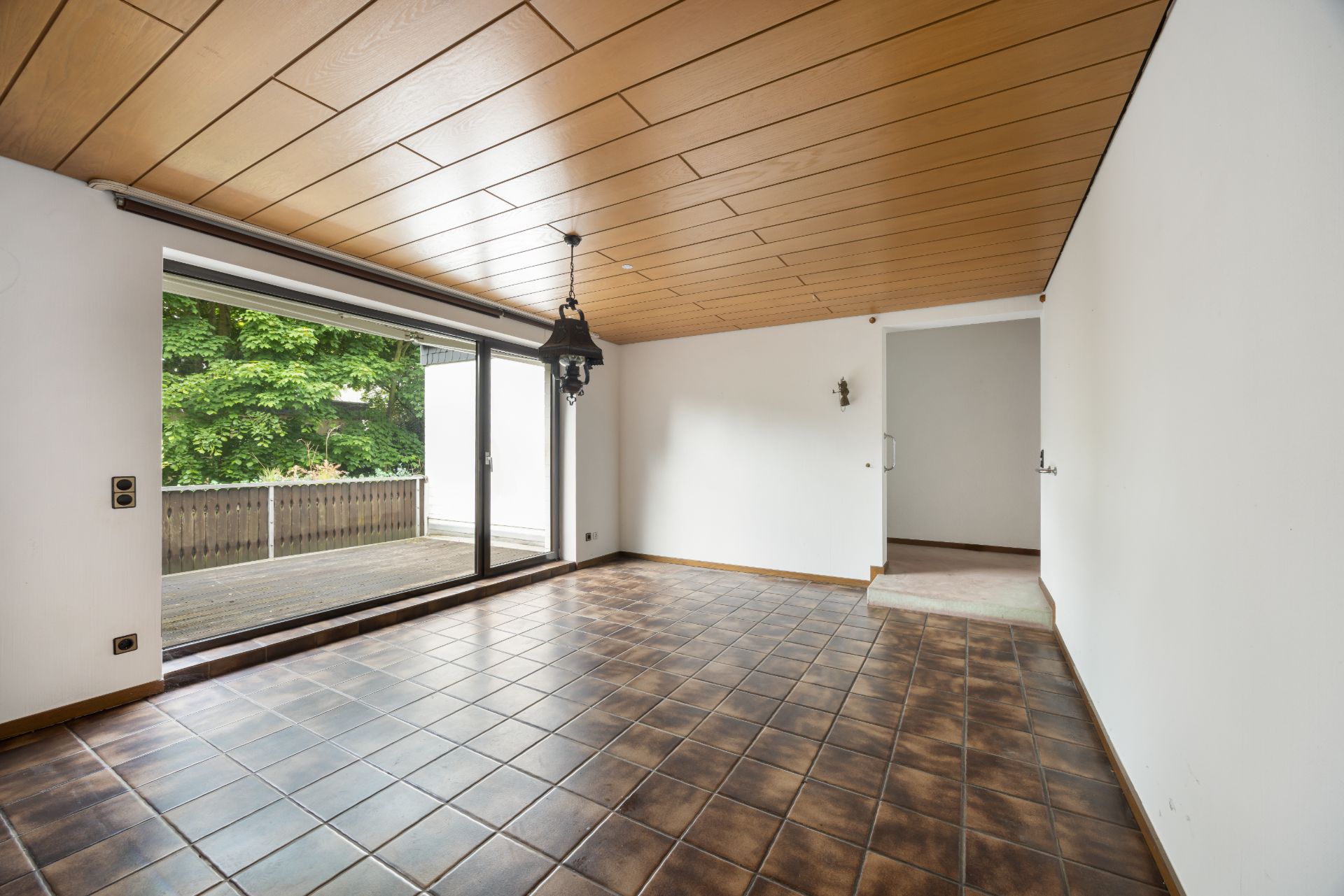 Leeres Zimmer mit braunen Fliesen, Holzdecke und großer Glasschiebetür zur Terrasse.