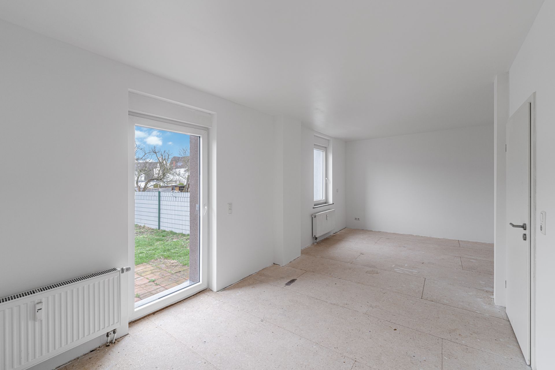 Leerer, heller Raum mit Tür zum Garten, zwei Fenstern und weißer Wand.