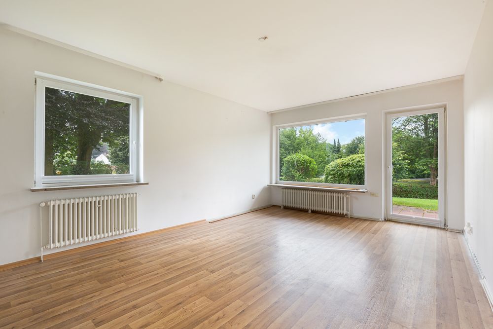 Leeres Wohnzimmer mit großen Fenstern, Holzboden und Blick auf den Garten.