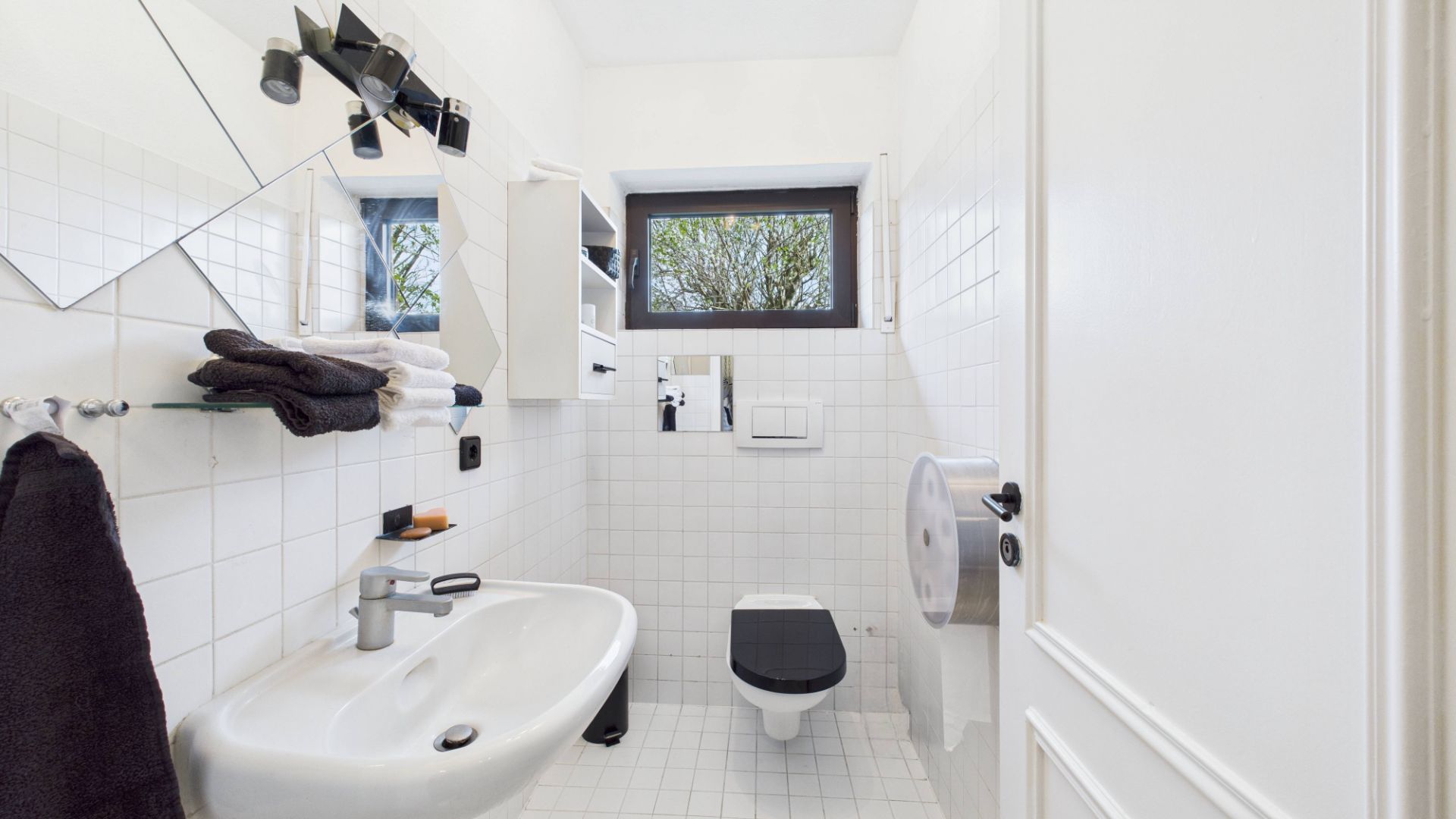 Helles Badezimmer mit Waschbecken, WC, Spiegel, Handtüchern und Fenster.