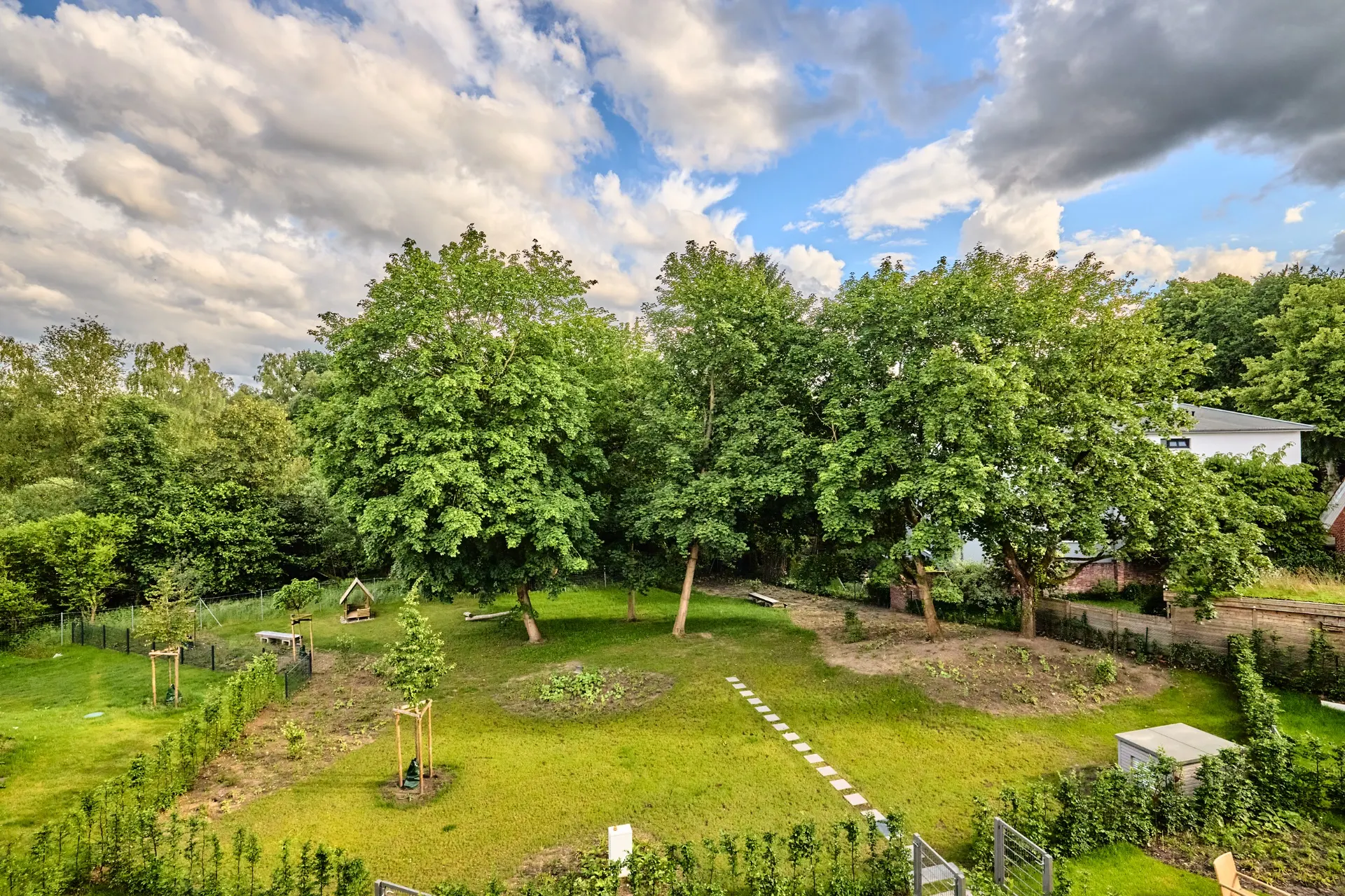 Garten mit mehreren großen Bäumen, Rasenfläche und einigen kleinen Beeten, im Hintergrund ein Haus.