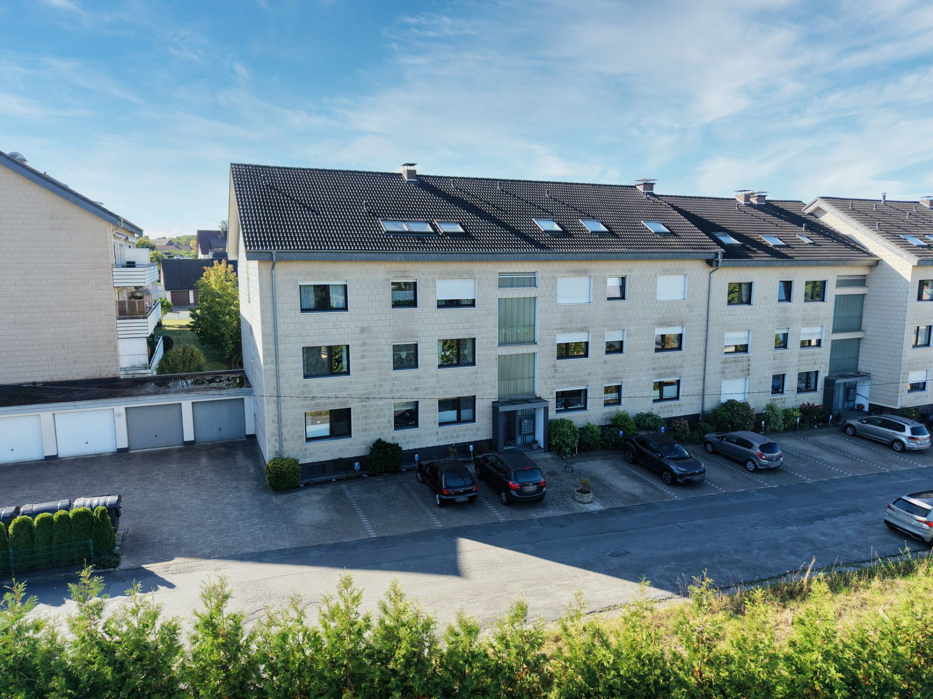 Mehrfamilienhaus mit drei Etagen, mehreren geparkten Autos und Garagen vor dem Gebäude.