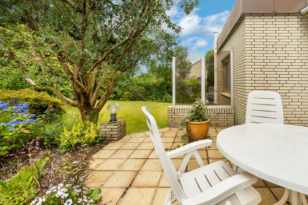 Gepflasterte Terrasse mit weißen Gartenmöbeln, Baum und blühenden Pflanzen im Garten.