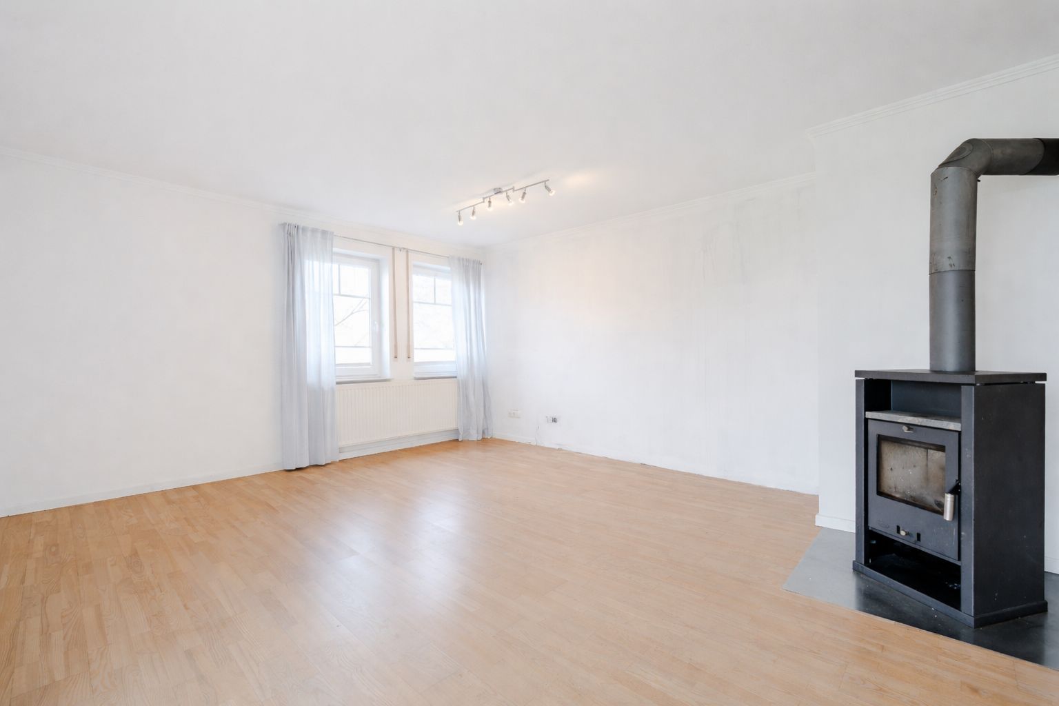 Helles Wohnzimmer mit Holzofen, zwei Fenstern und Parkettboden.