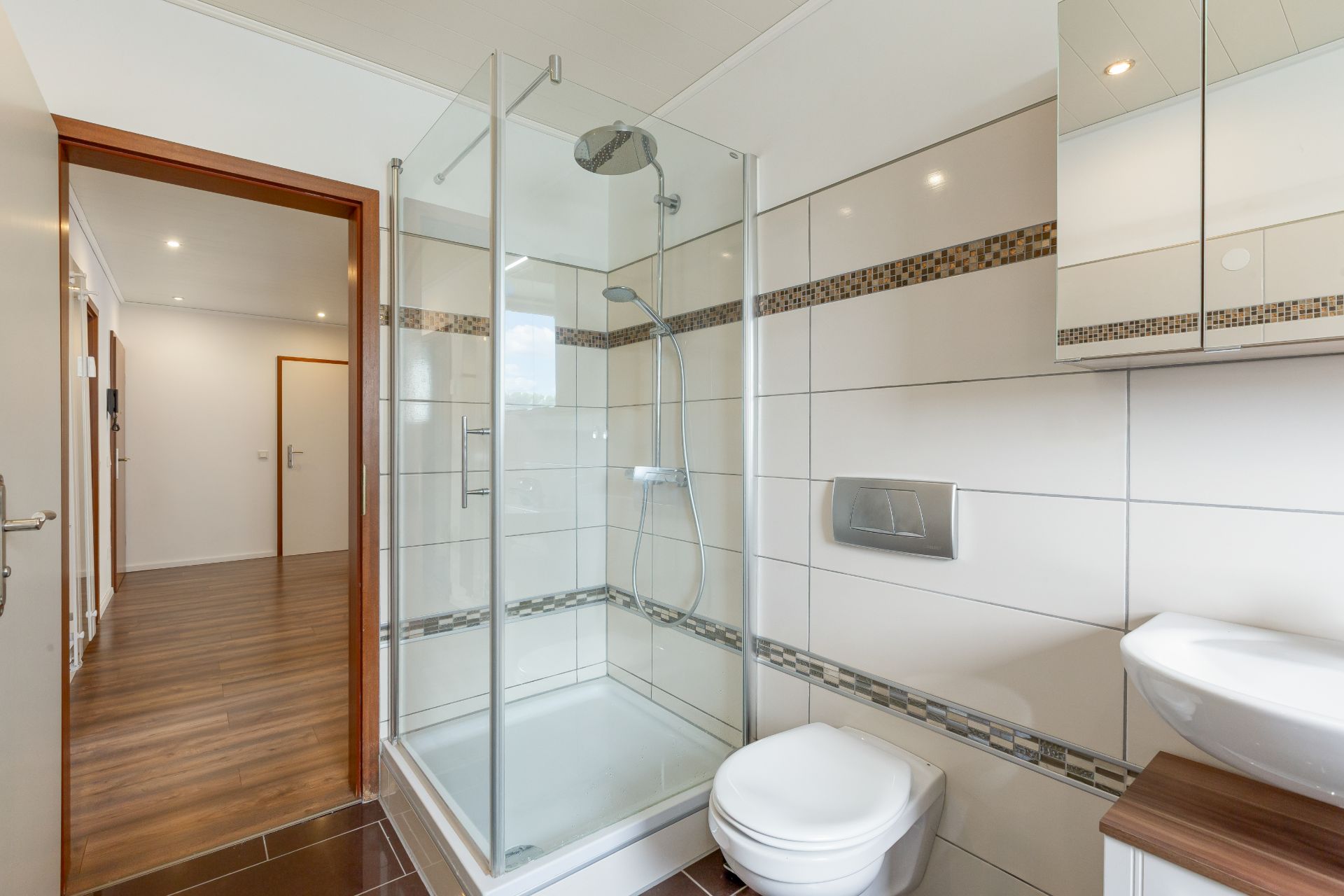Modernes Badezimmer mit Dusche, WC, Waschbecken und Fliesen, Tür zum Flur sichtbar.