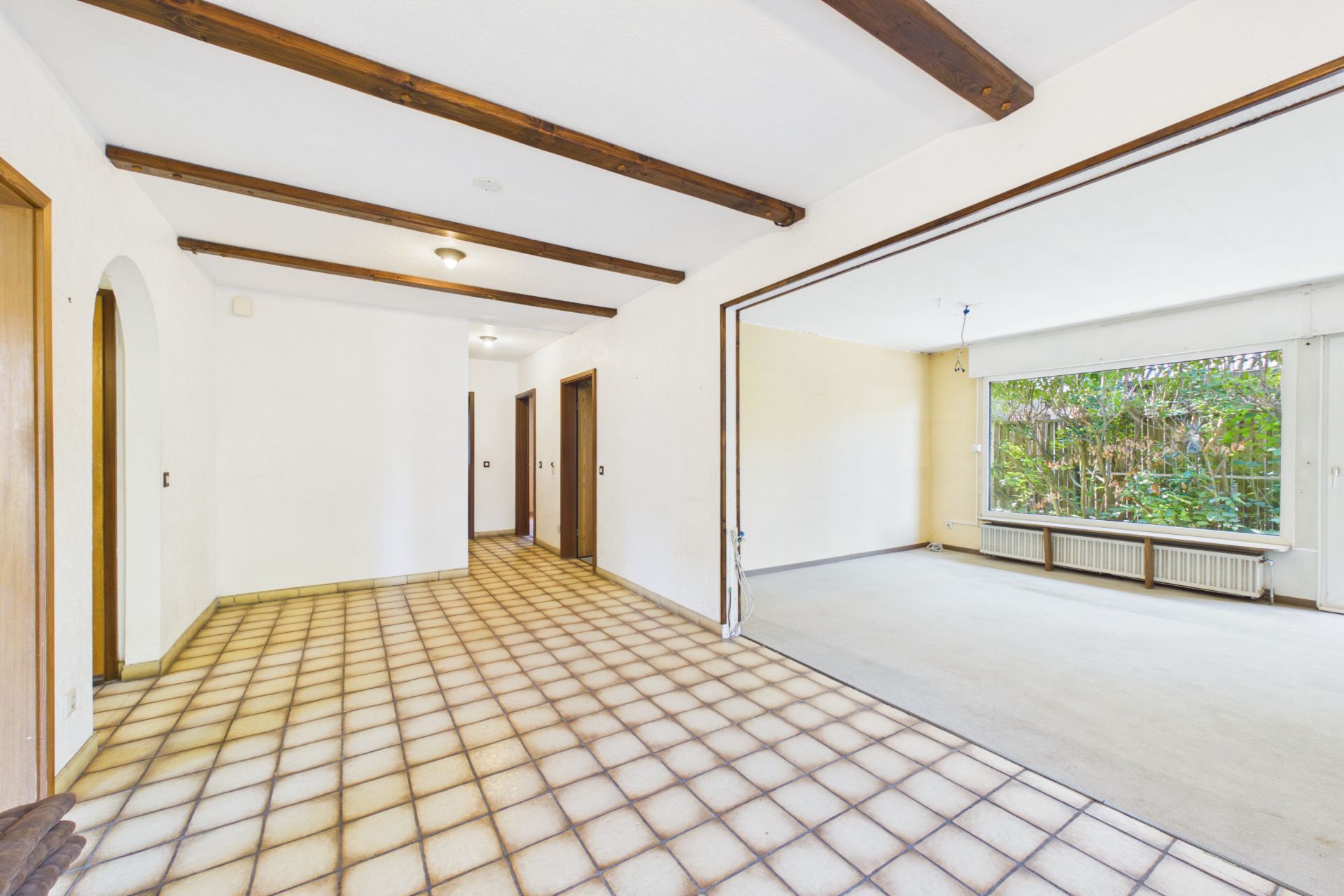 Helles Wohnzimmer mit Fliesenboden, Holzbalken und großem Fenster zum Garten.