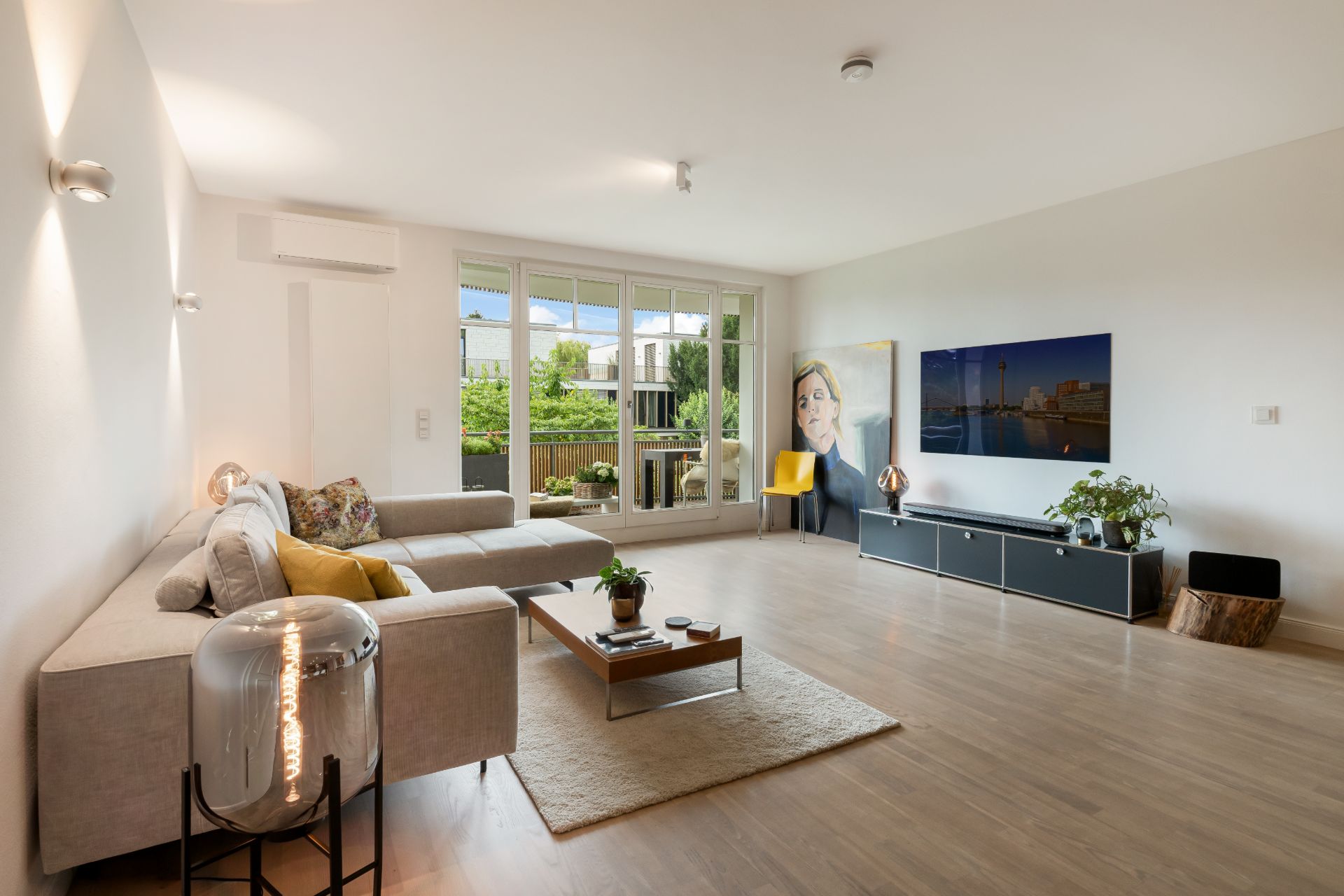 Helles Wohnzimmer mit Ecksofa, Couchtisch, Fernseher, Bildern und Zugang zum Balkon.