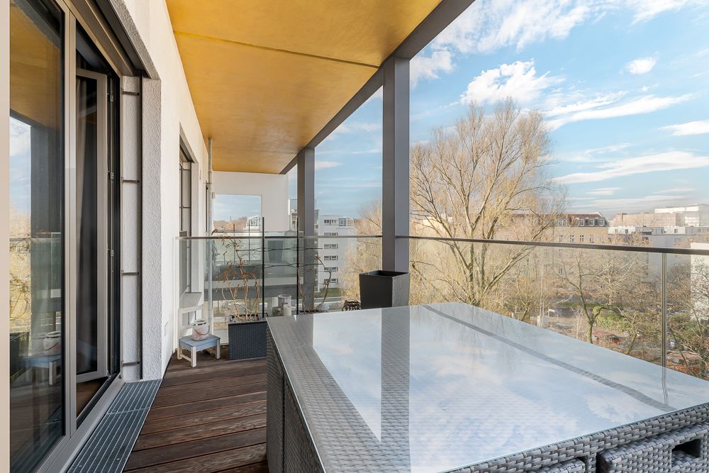 Großer Balkon mit Glasgeländer, Holzboden, Tisch und Blick auf Bäume und Stadt.