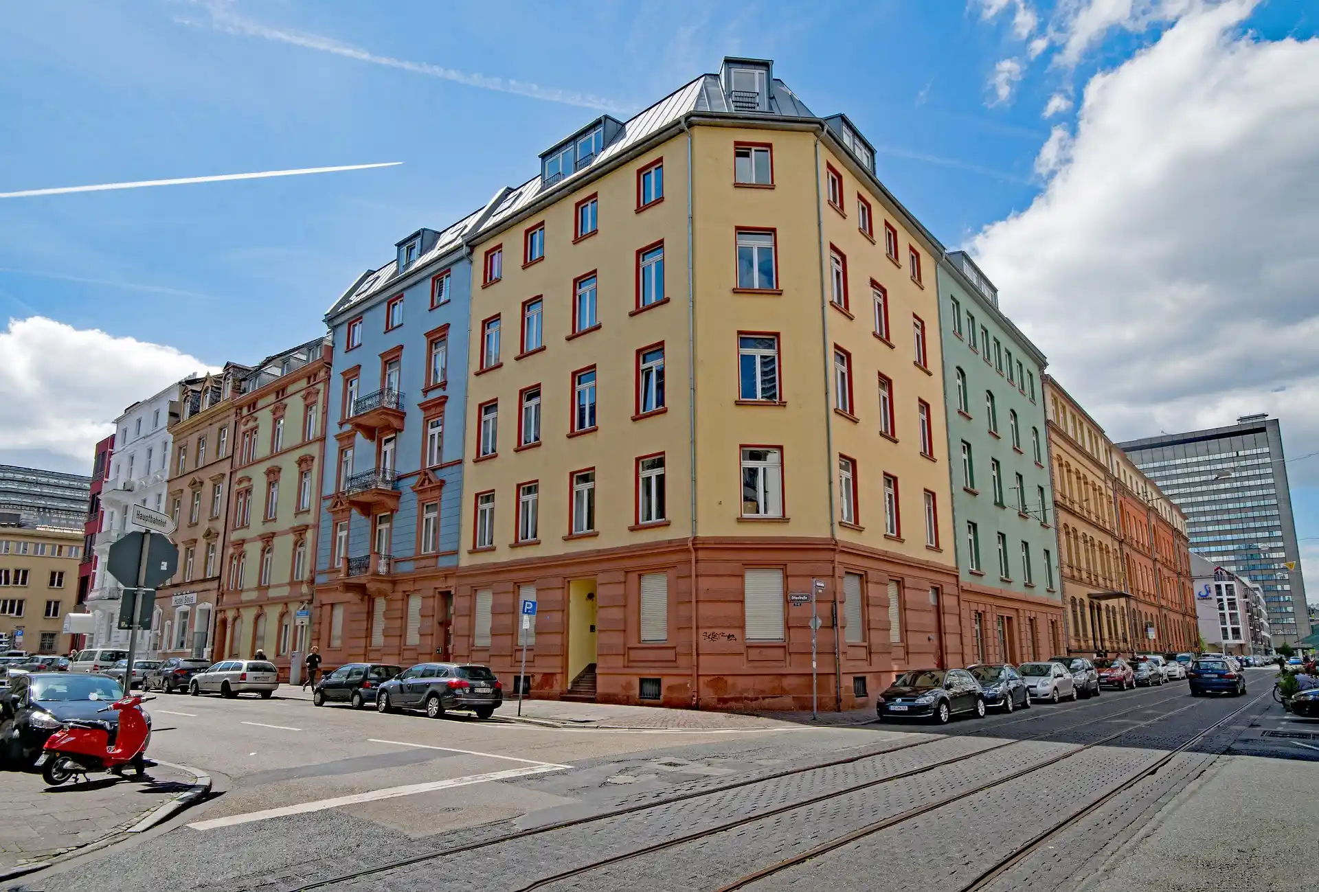Mehrstöckiges Wohnhaus mit bunten Fassaden an einer Straßenecke, Autos und Roller am Straßenrand.