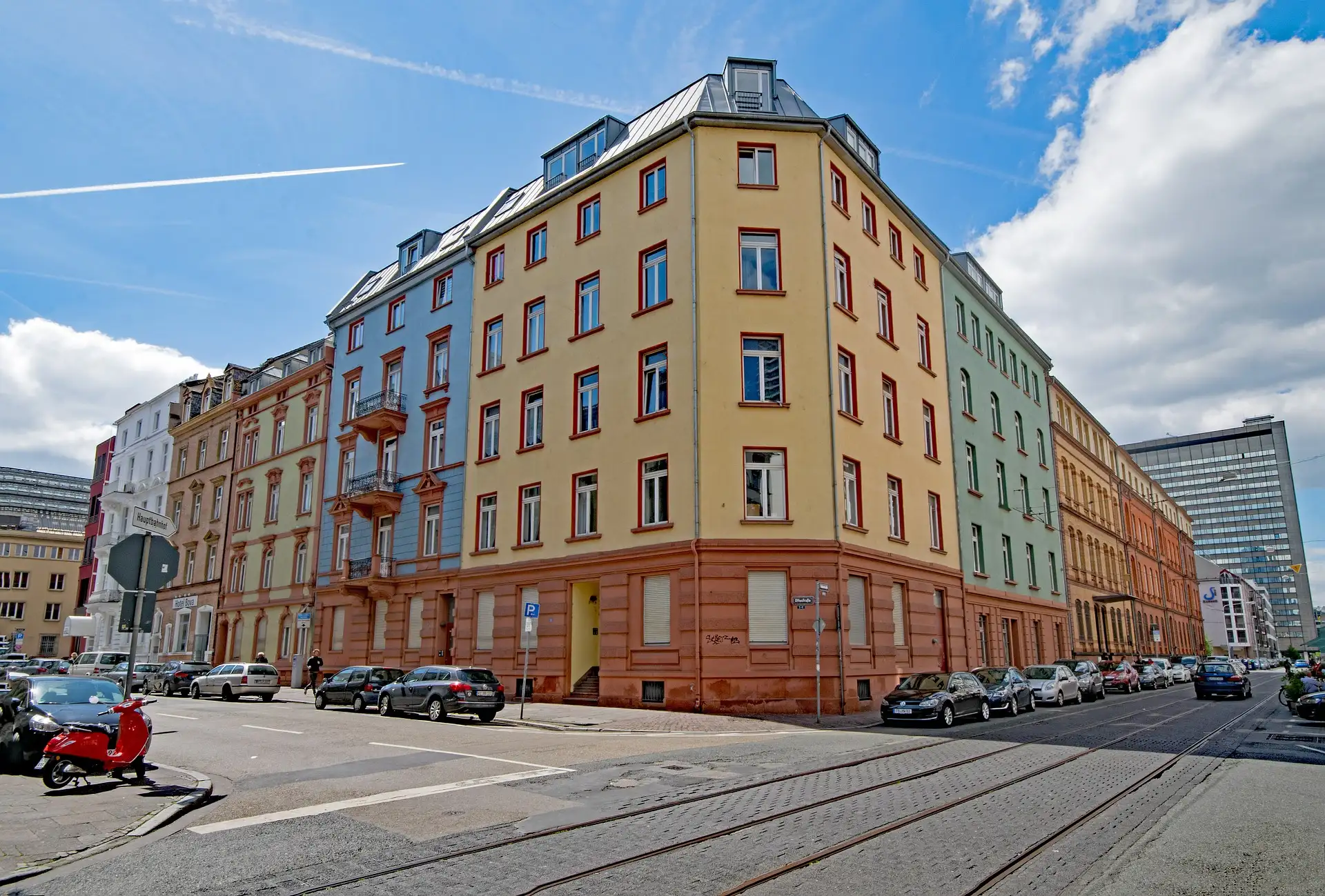 Mehrstöckiges Wohnhaus mit bunten Fassaden an einer Straßenecke, Autos und Roller am Straßenrand.