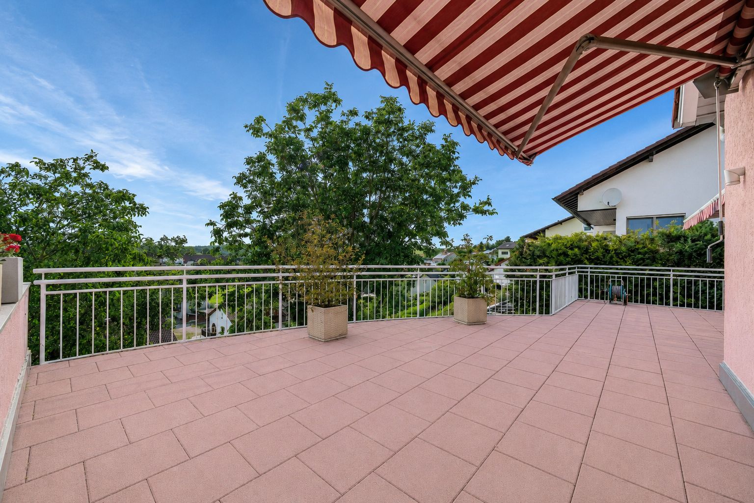 Große Terrasse mit rot-weißer Markise, Topfpflanzen und Blick auf Bäume und Nachbarhaus.