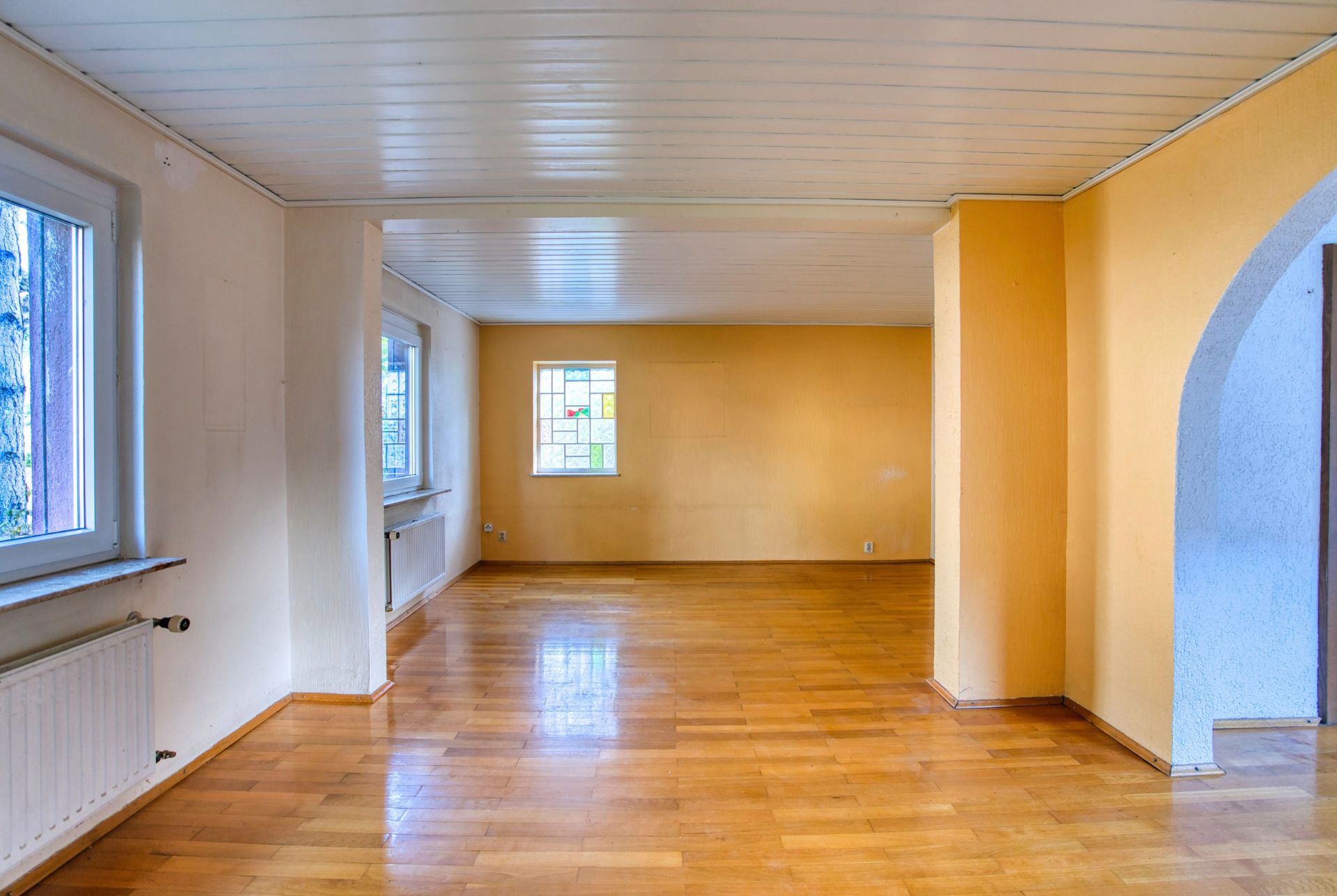 Helles, leeres Wohnzimmer mit Holzfußboden, gelben Wänden und Fenstern.