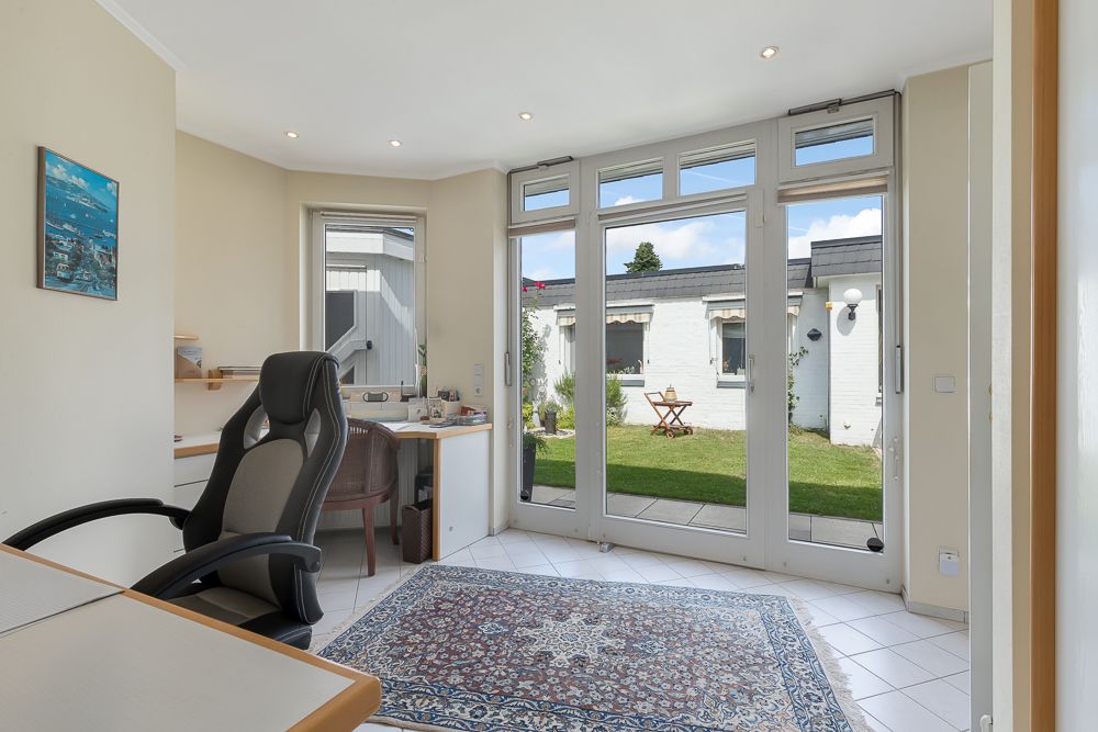 Helles Arbeitszimmer mit Schreibtisch, Bürostuhl, Teppich und Blick auf den Garten durch große Fenster.