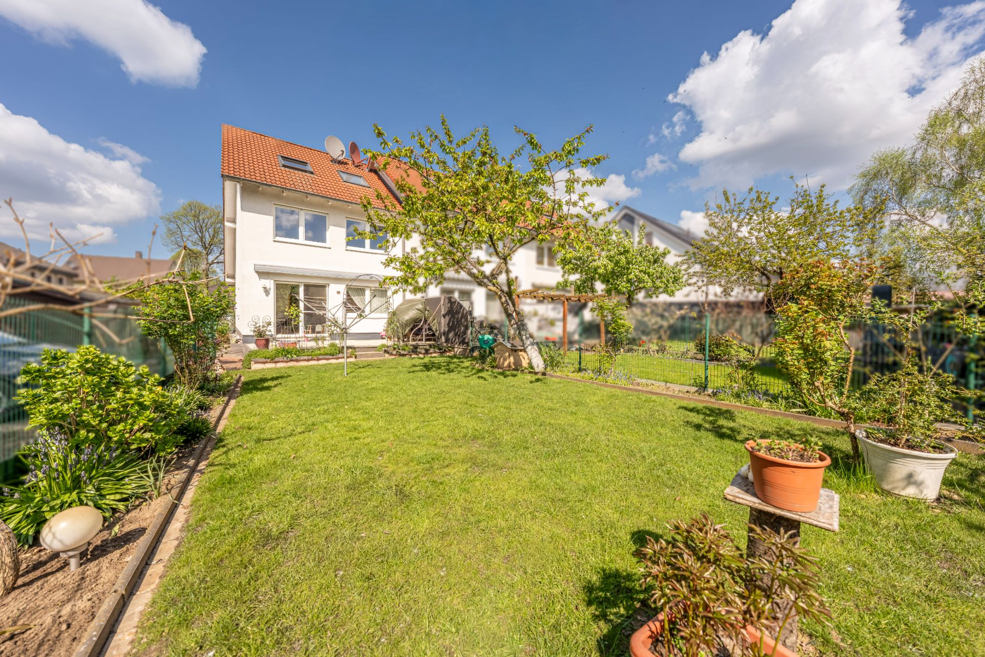 Grüner Garten mit Rasen, Bäumen und Pflanzen hinter einem weißen Wohnhaus an einem sonnigen Tag.