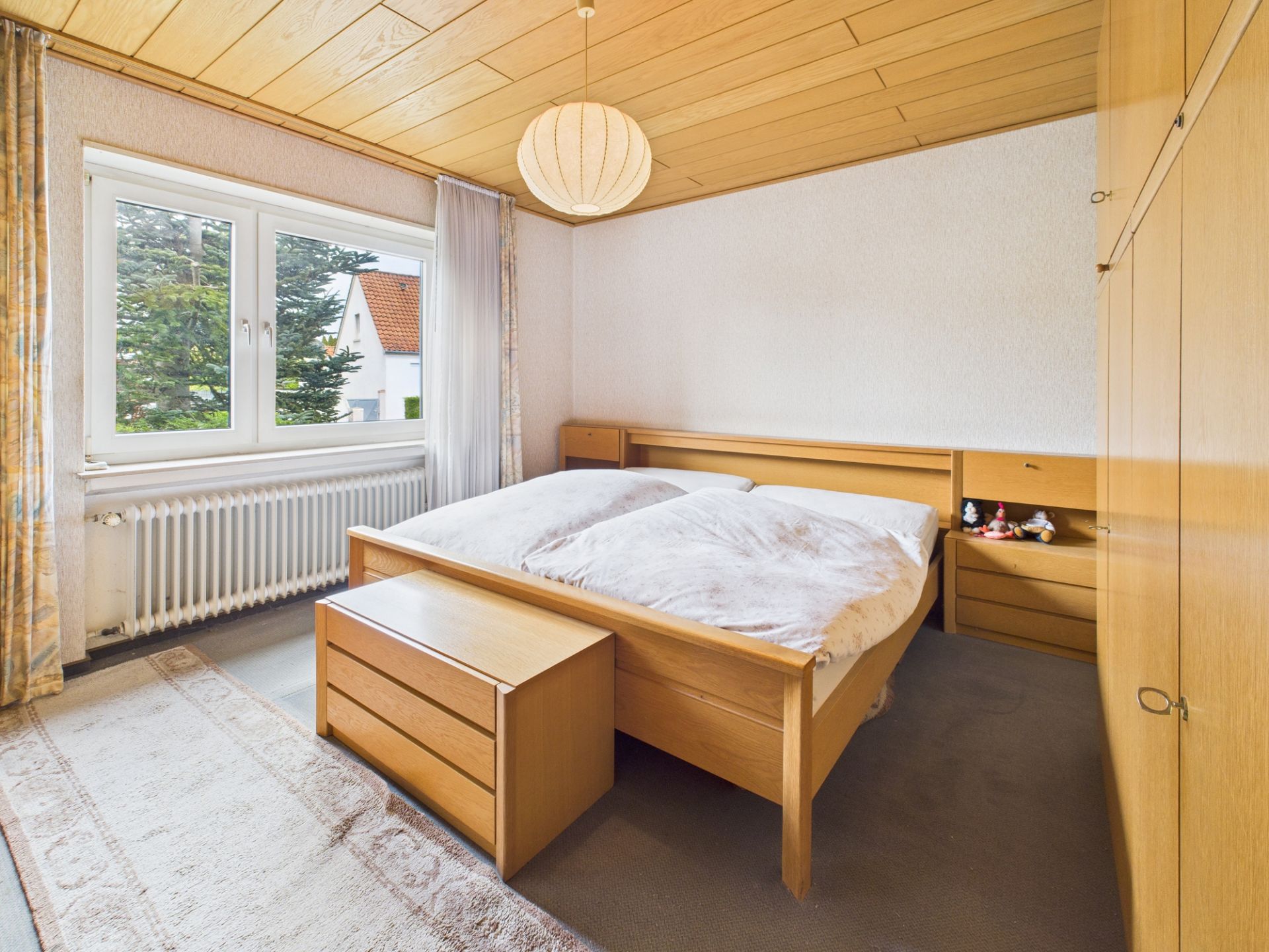 Helles Schlafzimmer mit Doppelbett aus Holz, großem Schrank und Fenster mit Blick nach draußen.