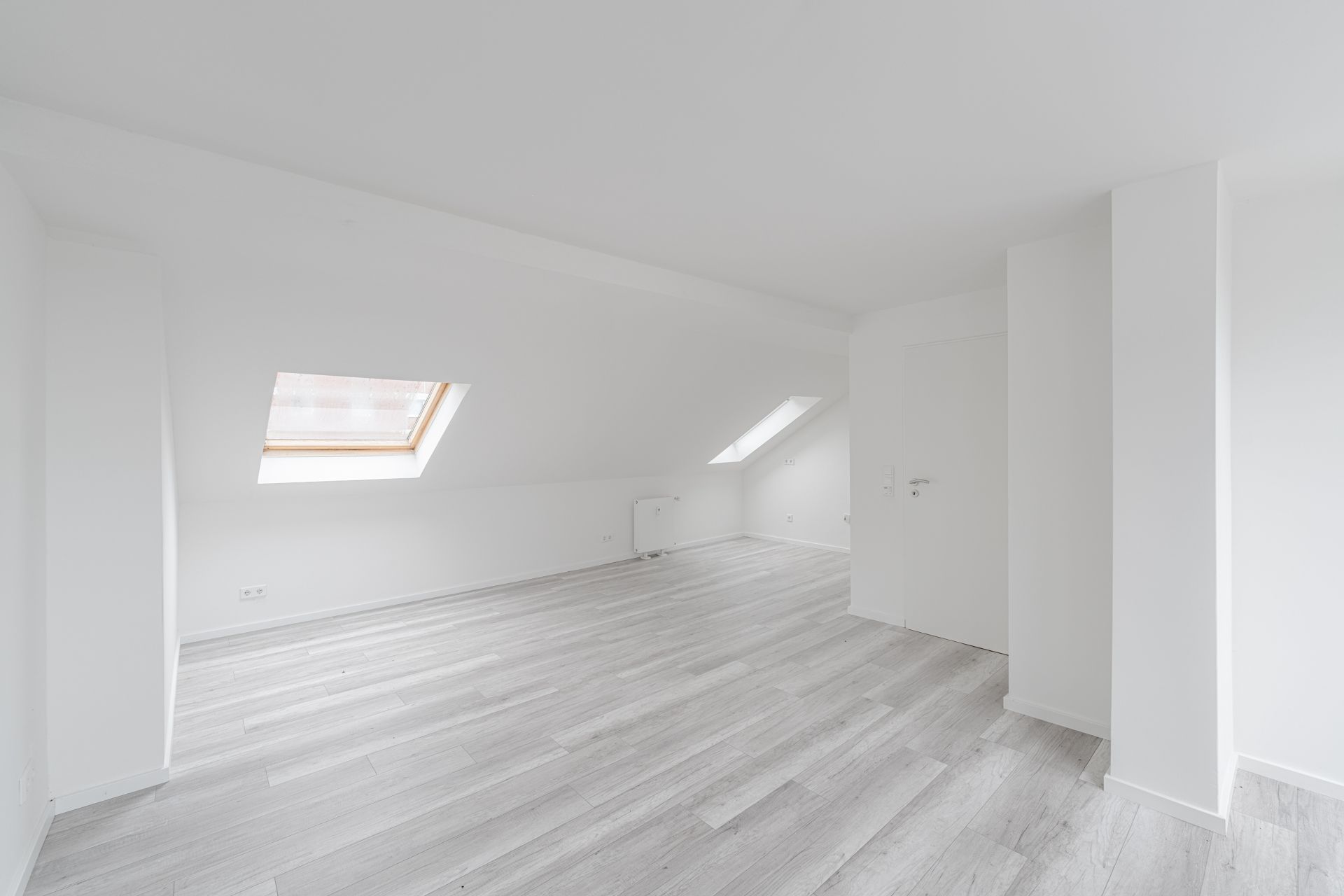Heller Dachraum mit zwei Dachfenstern, weißen Wänden und hellem Holzfußboden.