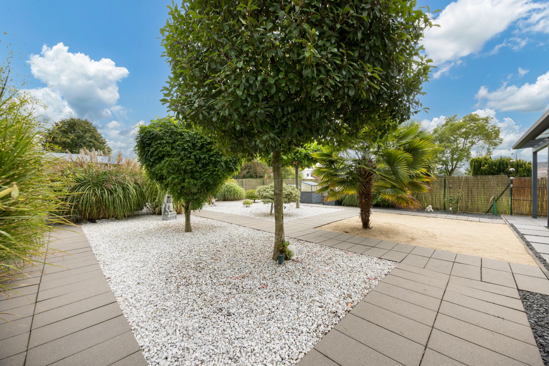 Garten mit Kiesflächen, Bäumen und gepflasterten Wegen bei blauem Himmel.