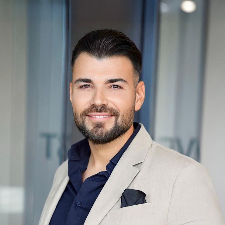 Mann mit Bart in hellem Anzug und dunklem Hemd steht vor Glaswand.