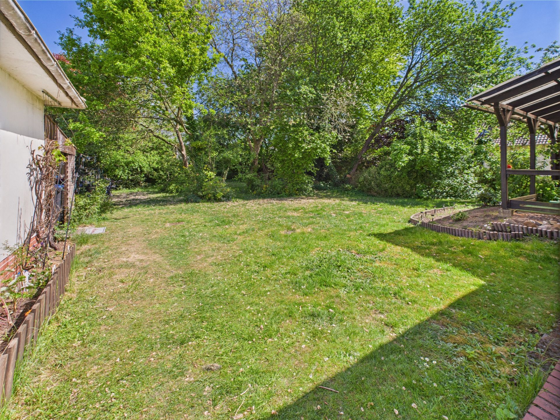 Grüner Garten mit Rasen, Bäumen und überdachter Terrasse am rechten Rand.
