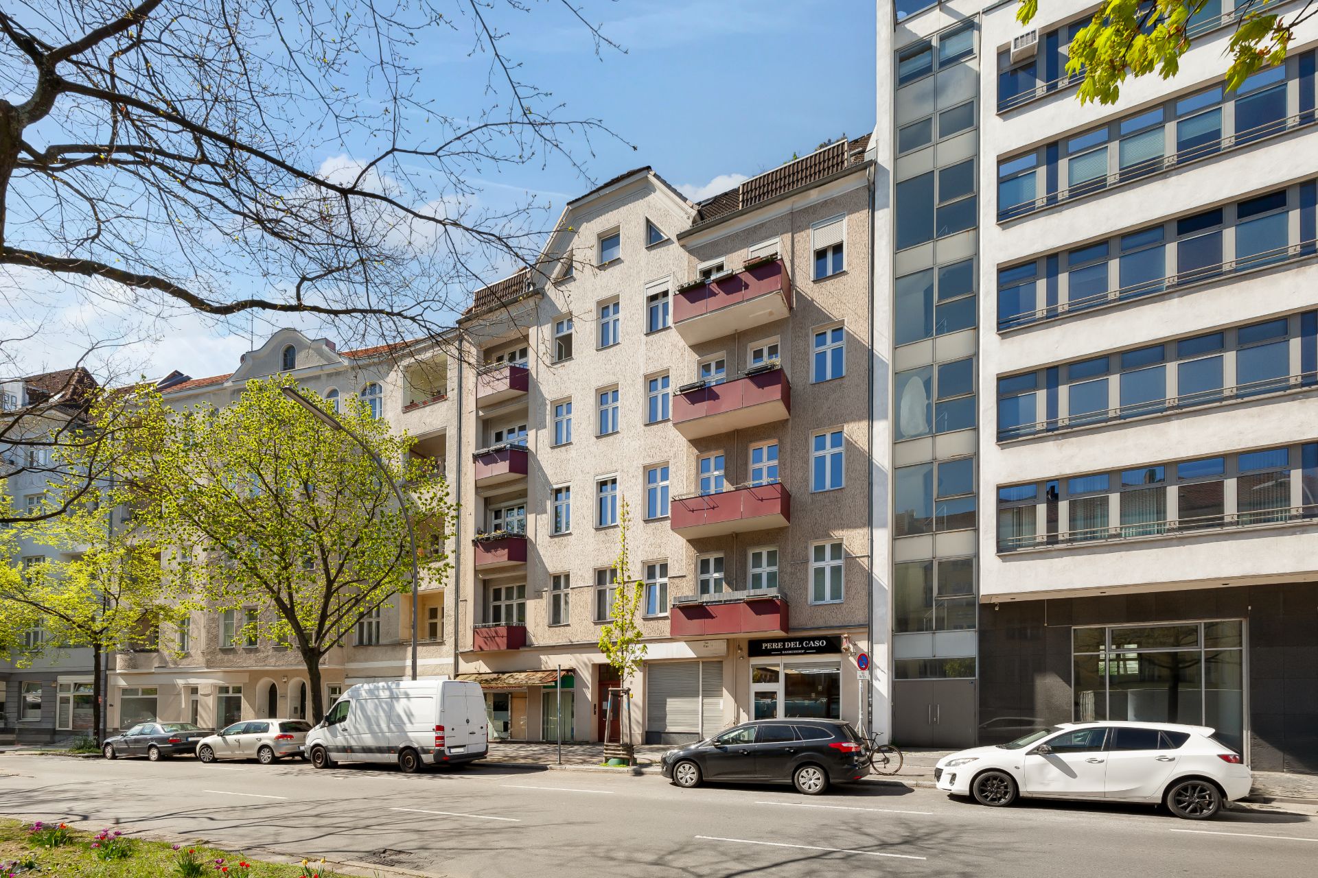 Mehrstöckiges Wohnhaus mit roten Balkonen an einer Straße, mehrere geparkte Autos davor.