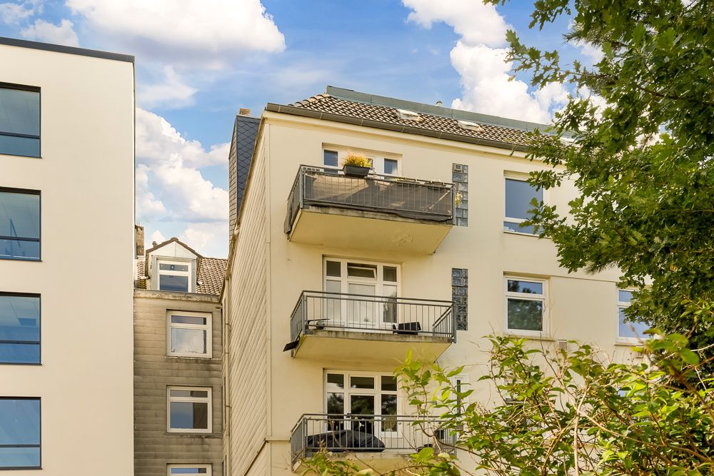 Mehrfamilienhaus mit drei Balkonen, hellem Putz und umgeben von Bäumen.