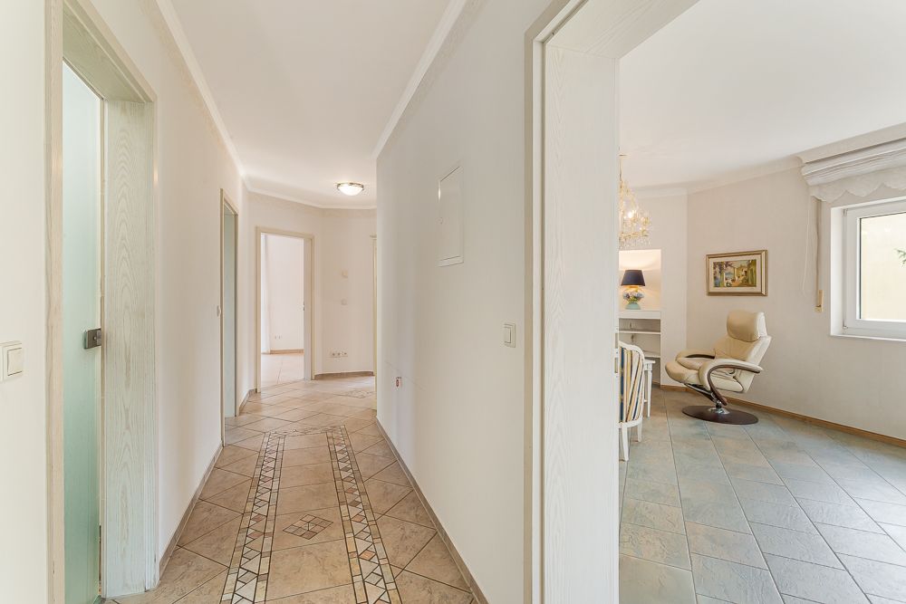 Heller Flur mit Fliesenboden, Zugang zu Zimmern, Wohnzimmer mit Sessel und Fenster sichtbar.