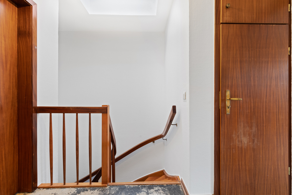 Innenraum mit Holztreppengeländer, weißer Wand und Holztür auf der rechten Seite.