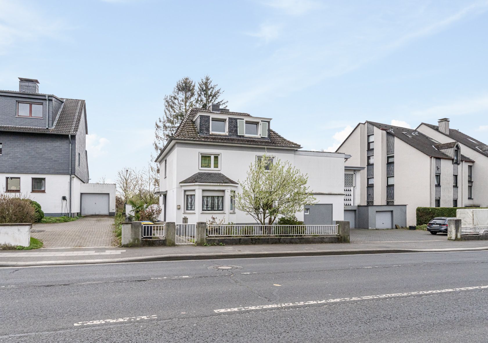 Zweistöckiges weißes Wohnhaus mit Dachfenstern, Vorgarten und Einfahrt an einer Straße.