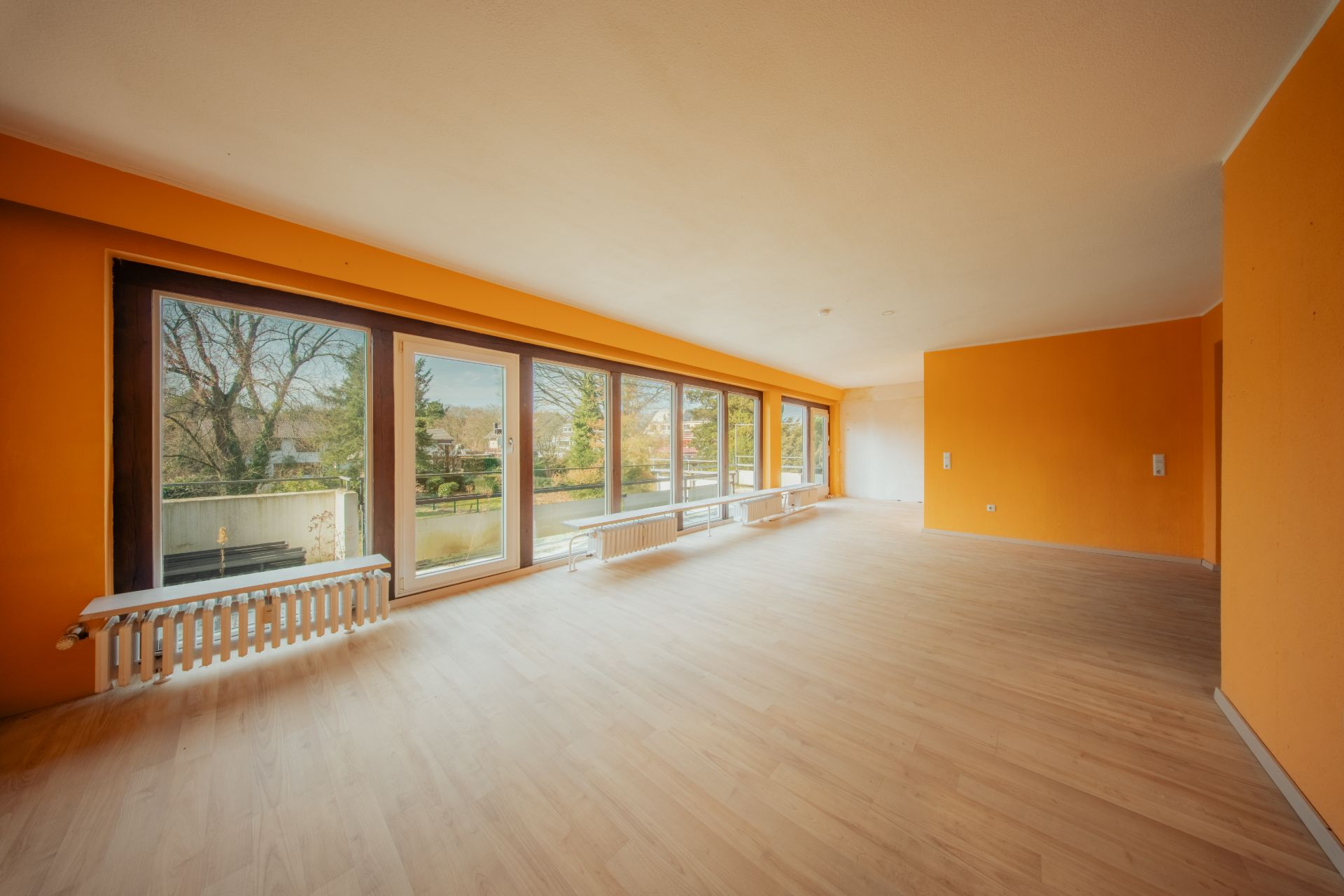 Großer, leerer Raum mit orangefarbenen Wänden und großen Fenstern mit Blick auf Garten.