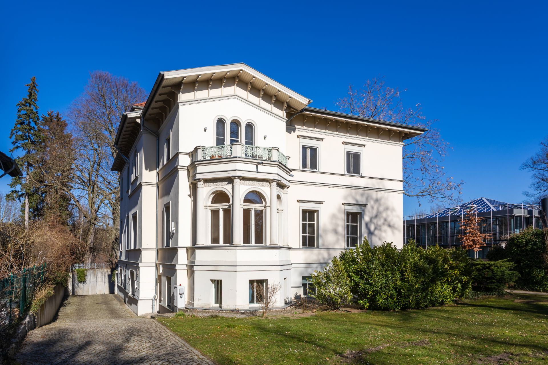 Großes, weißes Wohnhaus mit Erker, Garten und gepflasterter Zufahrt bei blauem Himmel.