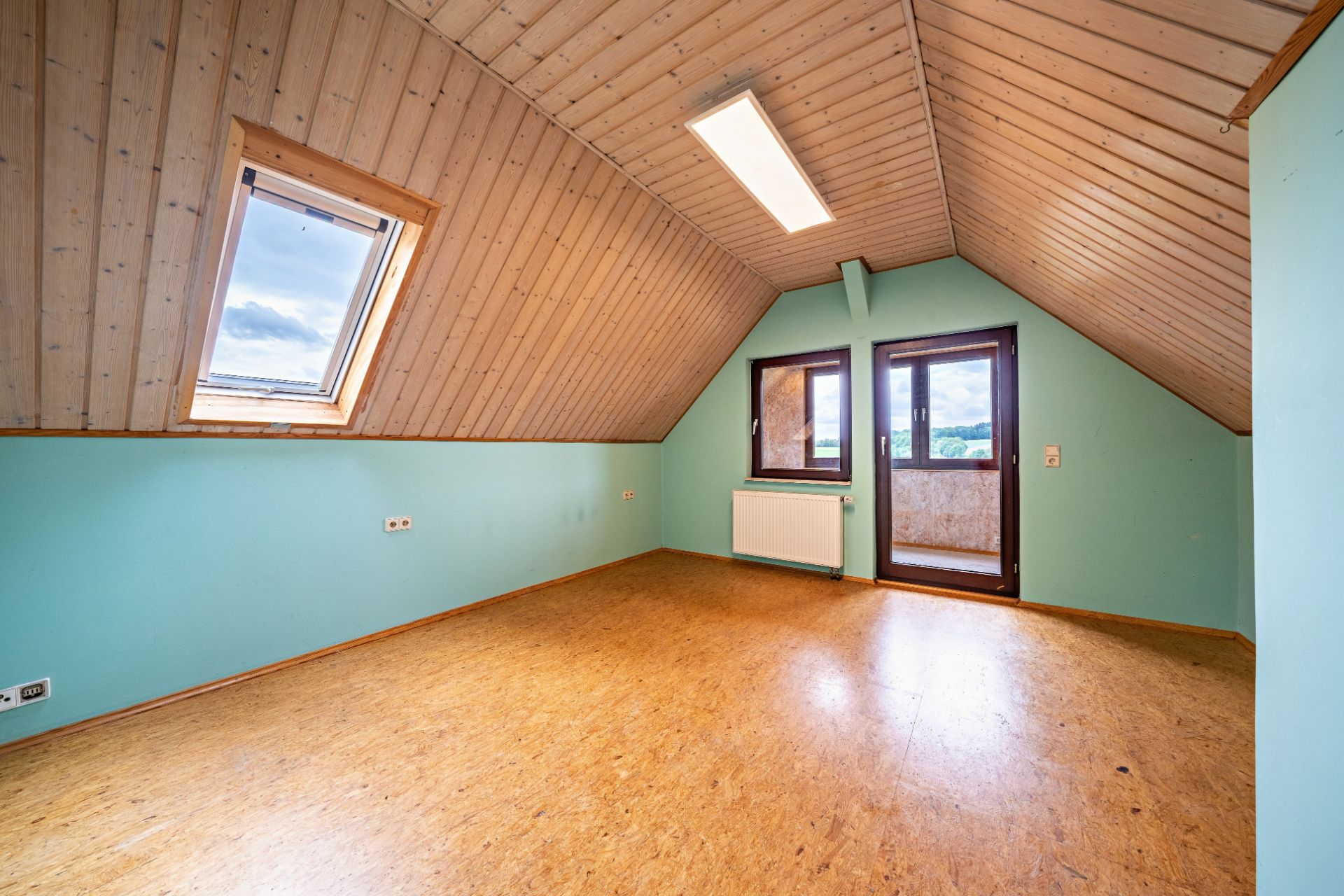Dachzimmer mit Holzdecke, Fenster, Tür zum Balkon und grün gestrichenen Wänden.