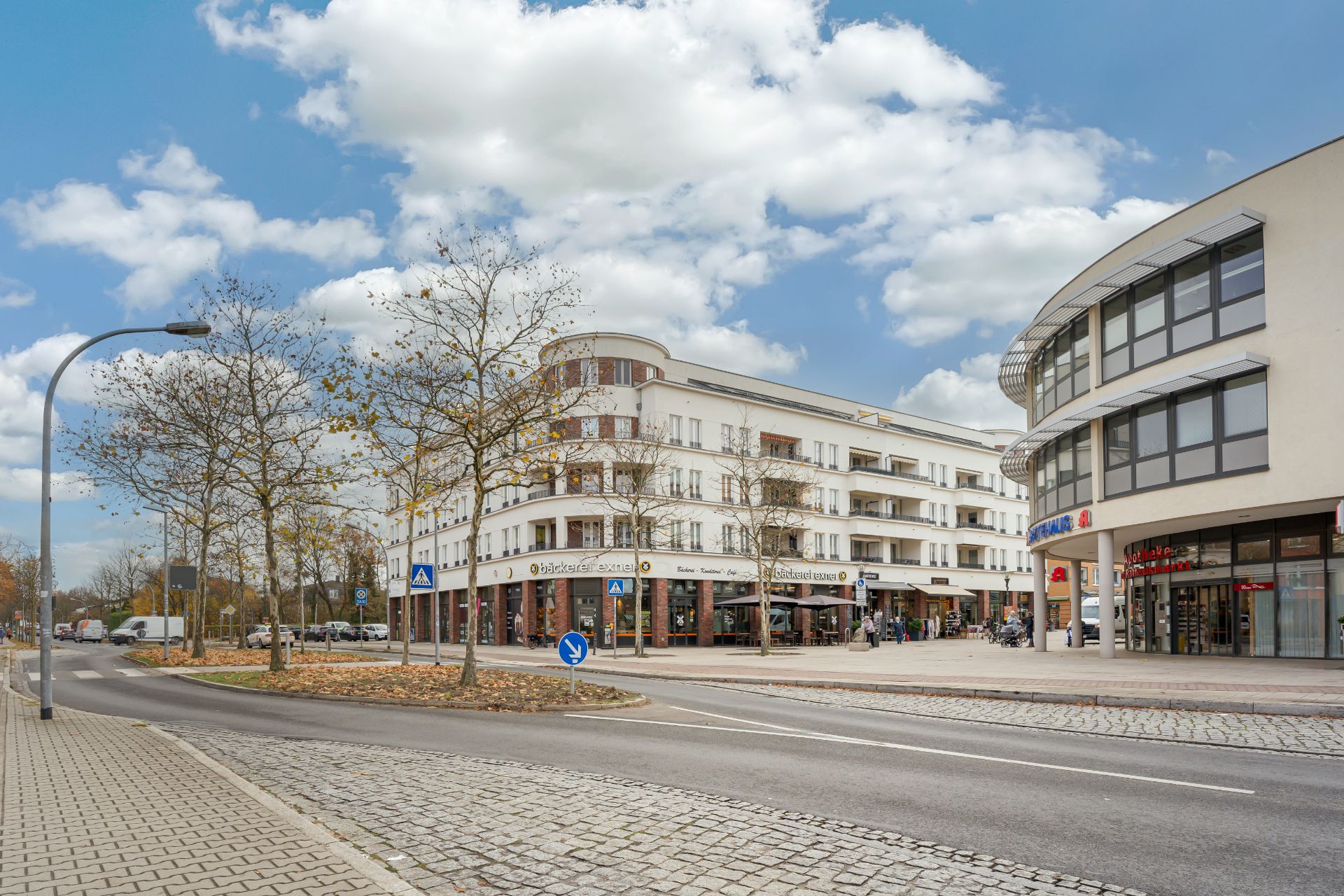 Mehrstöckiges Wohngebäude mit Balkonen an einer Straßenkreuzung, Geschäfte im Erdgeschoss.