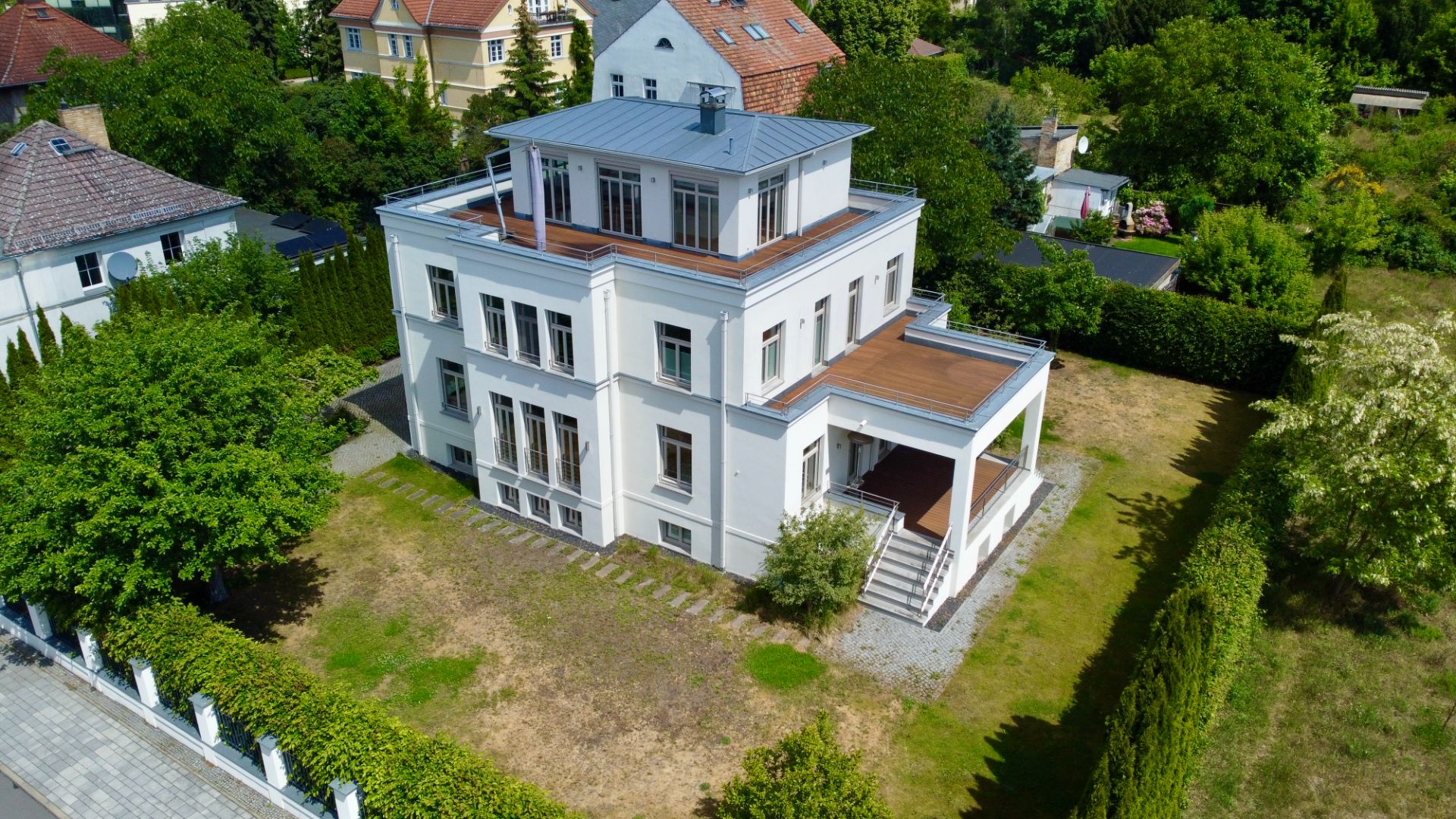 Großes weißes Wohnhaus mit mehreren Etagen und Flachdach, umgeben von Garten und Bäumen.