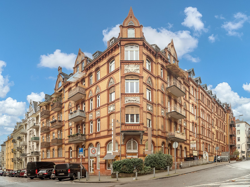 Mehrstöckiges Backsteinhaus mit Balkonen an einer Straßenecke, Autos parken am Straßenrand.