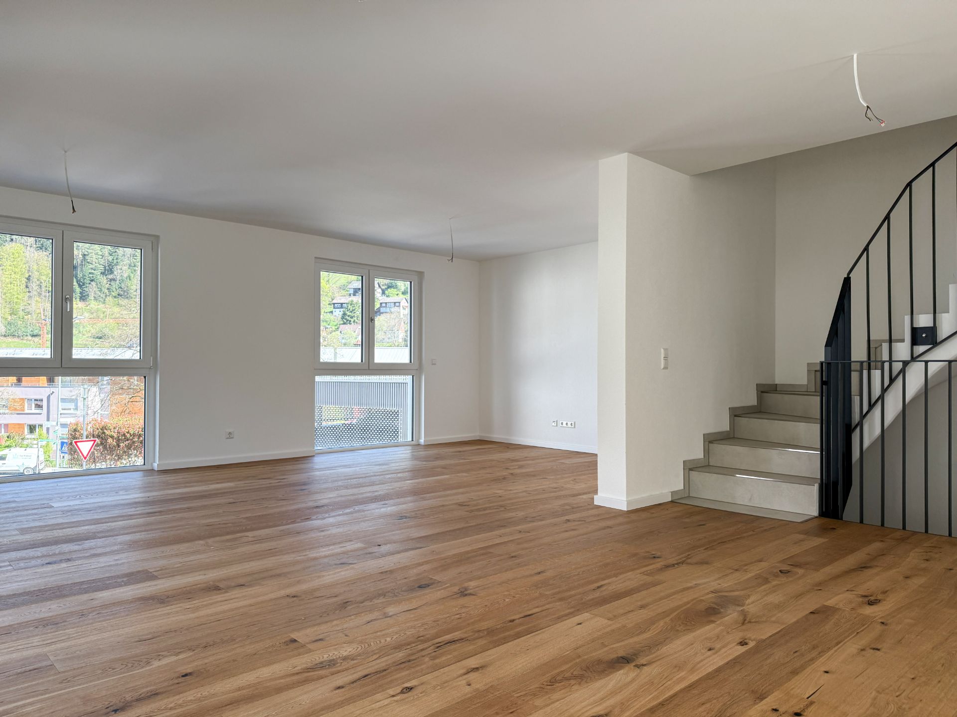 Helles Wohnzimmer mit Holzboden, großen Fenstern und Treppe mit schwarzem Geländer.