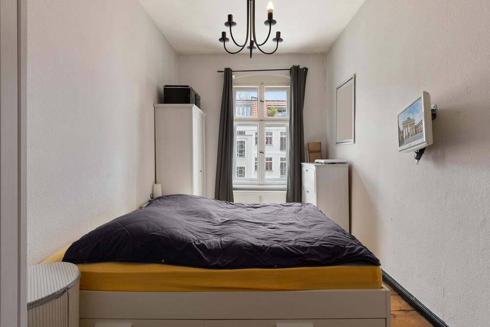 Schlafzimmer mit Doppelbett, Kleiderschrank, Fenster mit Vorhängen und an der Wand montiertem Fernseher.